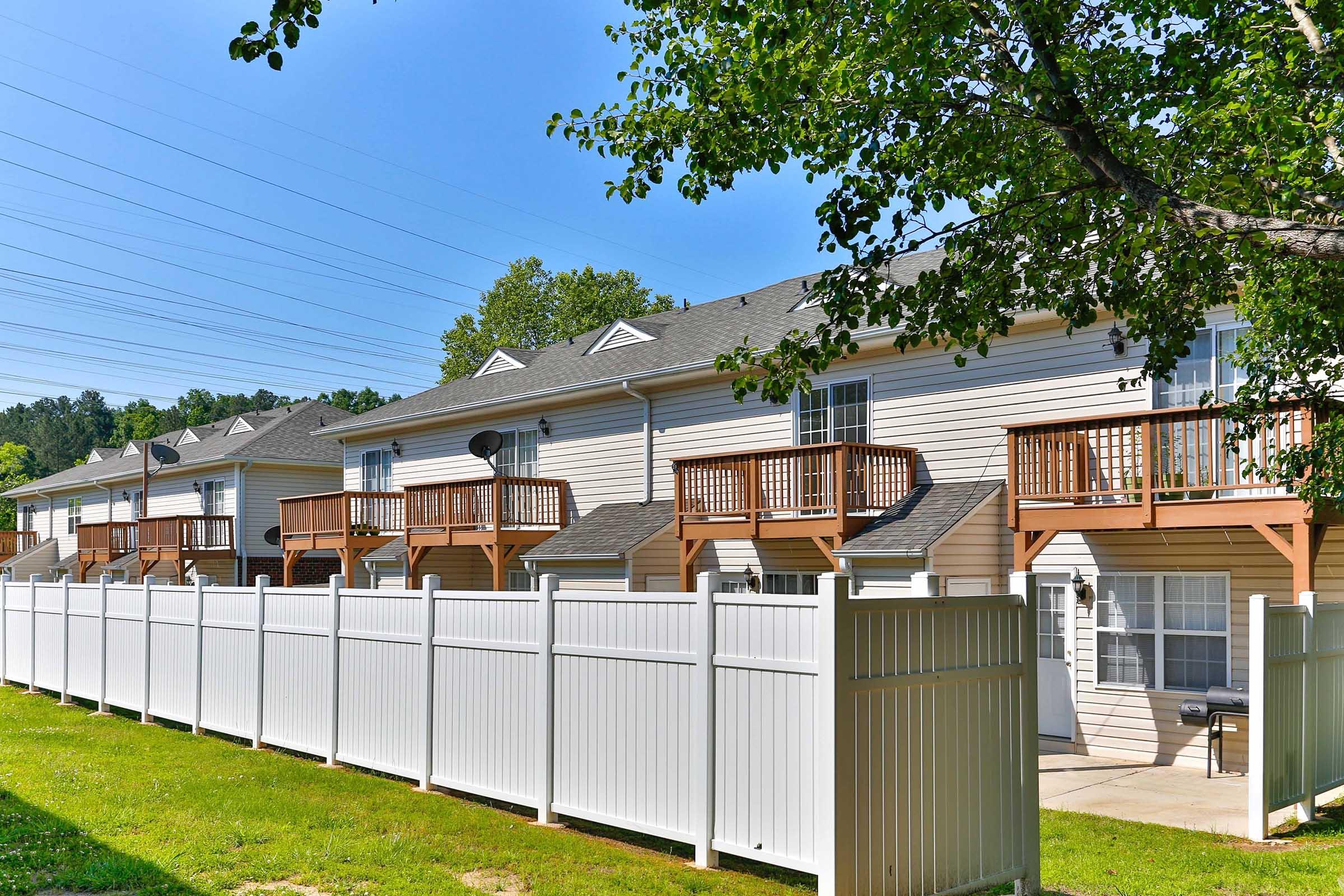 A row of townhouses with wooden decks, located behind a white privacy fence. The scene is set on a sunny day, with clear blue skies and greenery in the background. Power lines are visible against the sky, adding to the residential neighborhood's ambiance.