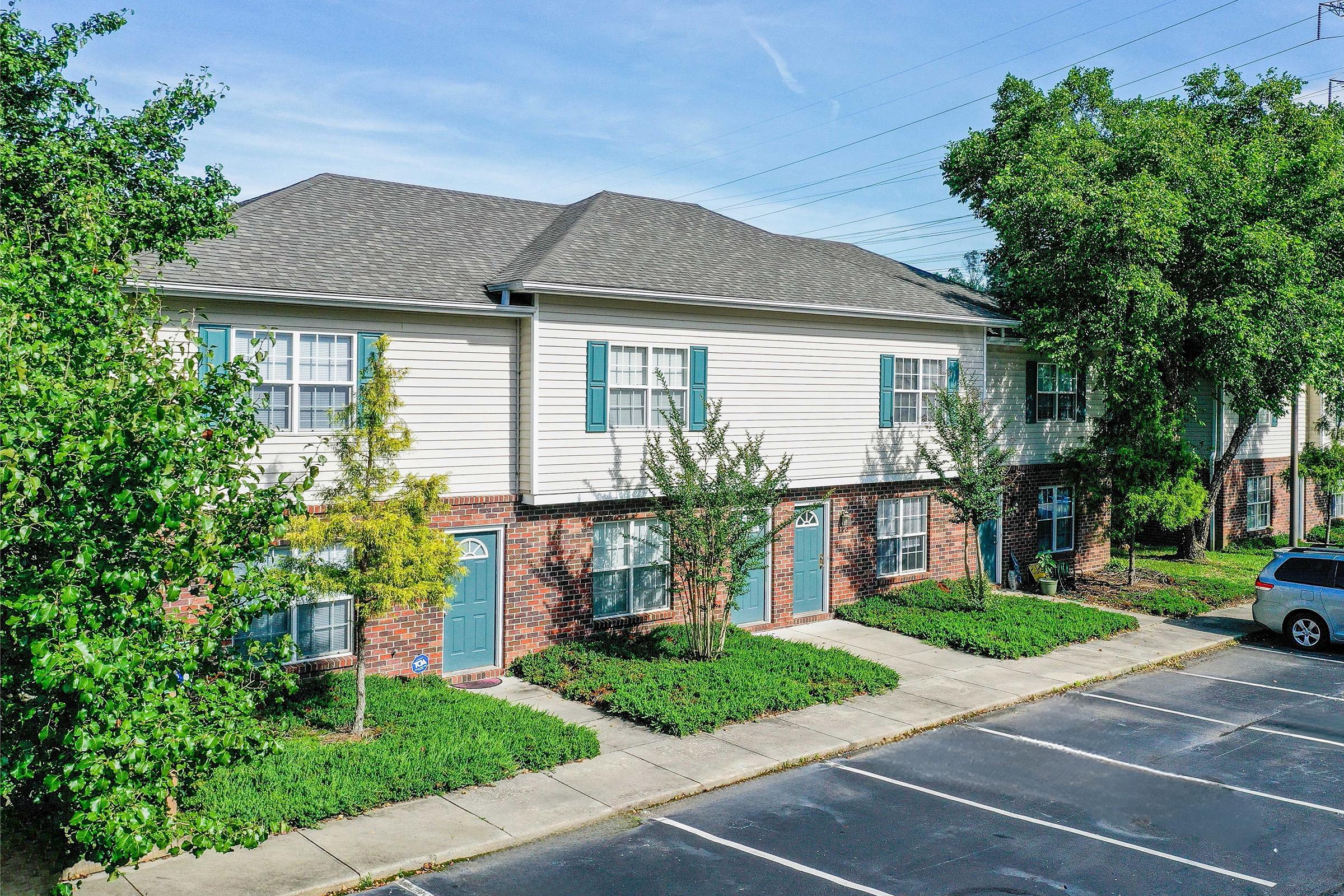 A two-story apartment building with a light-colored exterior and brick accents. The building has several shrubs and small trees in front, along with a parking lot featuring empty parking spaces. Clear blue sky in the background and visible power lines.