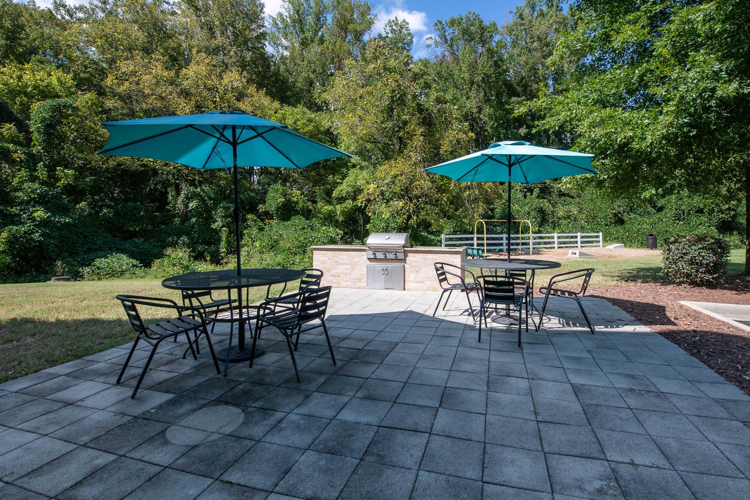 A spacious outdoor area featuring two turquoise umbrellas over black metal tables and chairs set on a stone patio. In the background, there is a grill and lush greenery, including trees and a fence, providing a serene environment for gatherings or relaxation.
