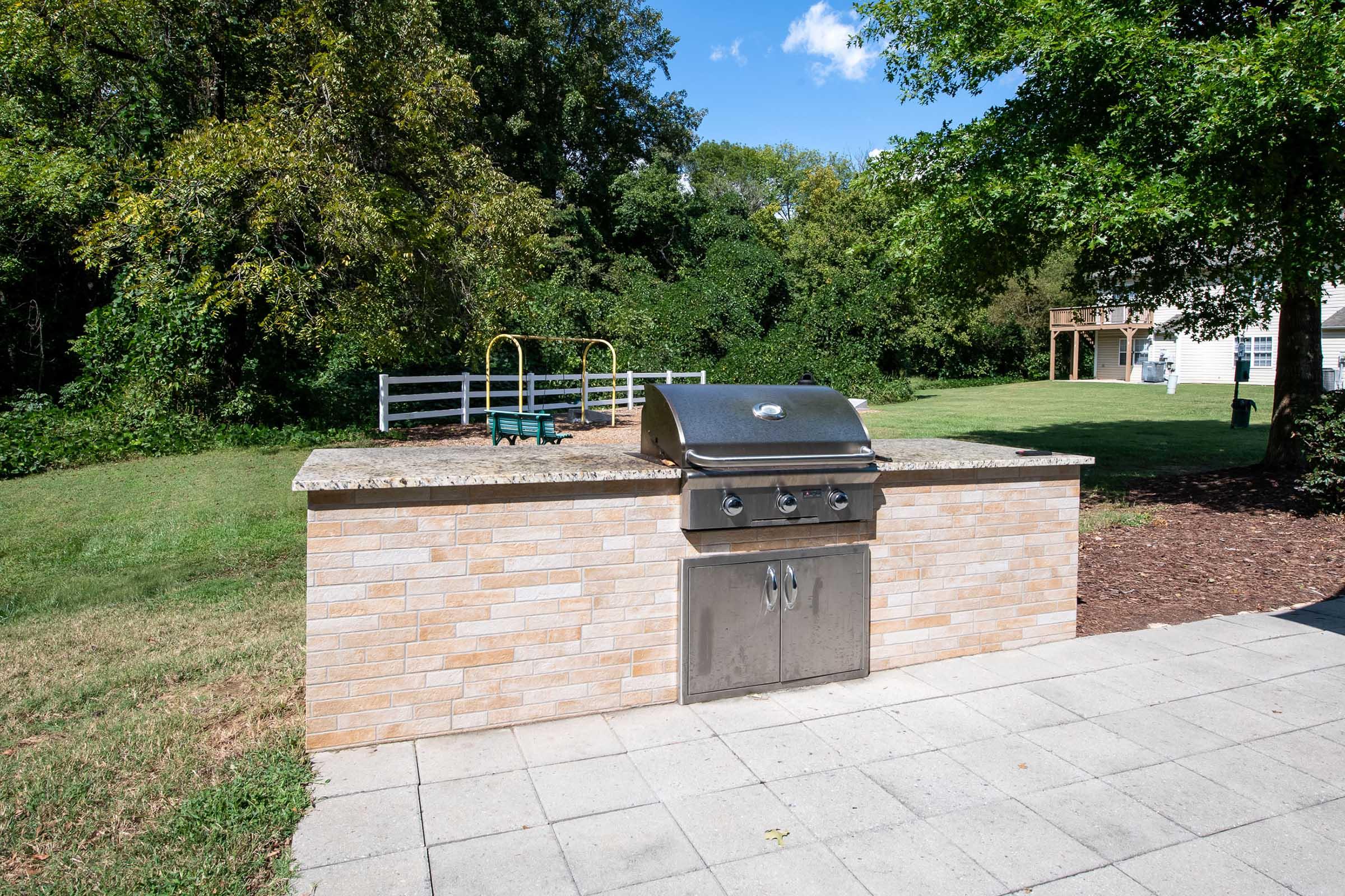 An outdoor barbecue grill set on a stone countertop, surrounded by a grassy yard. In the background, there is a playground with swings and a large tree, and a wooded area. The sky is clear with a few clouds, indicating a sunny day.