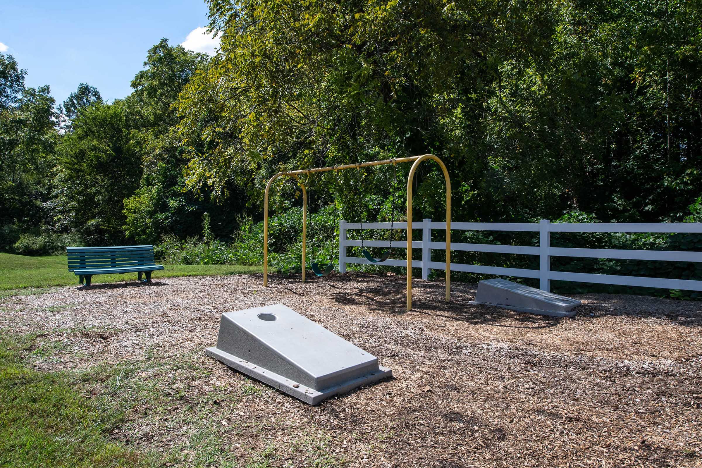 A playground area featuring a yellow swing set and a cornhole game on a bed of wood chips. In the background, there is a wooden fence and greenery, and a blue bench is positioned nearby, surrounded by a sunny, spacious park setting.