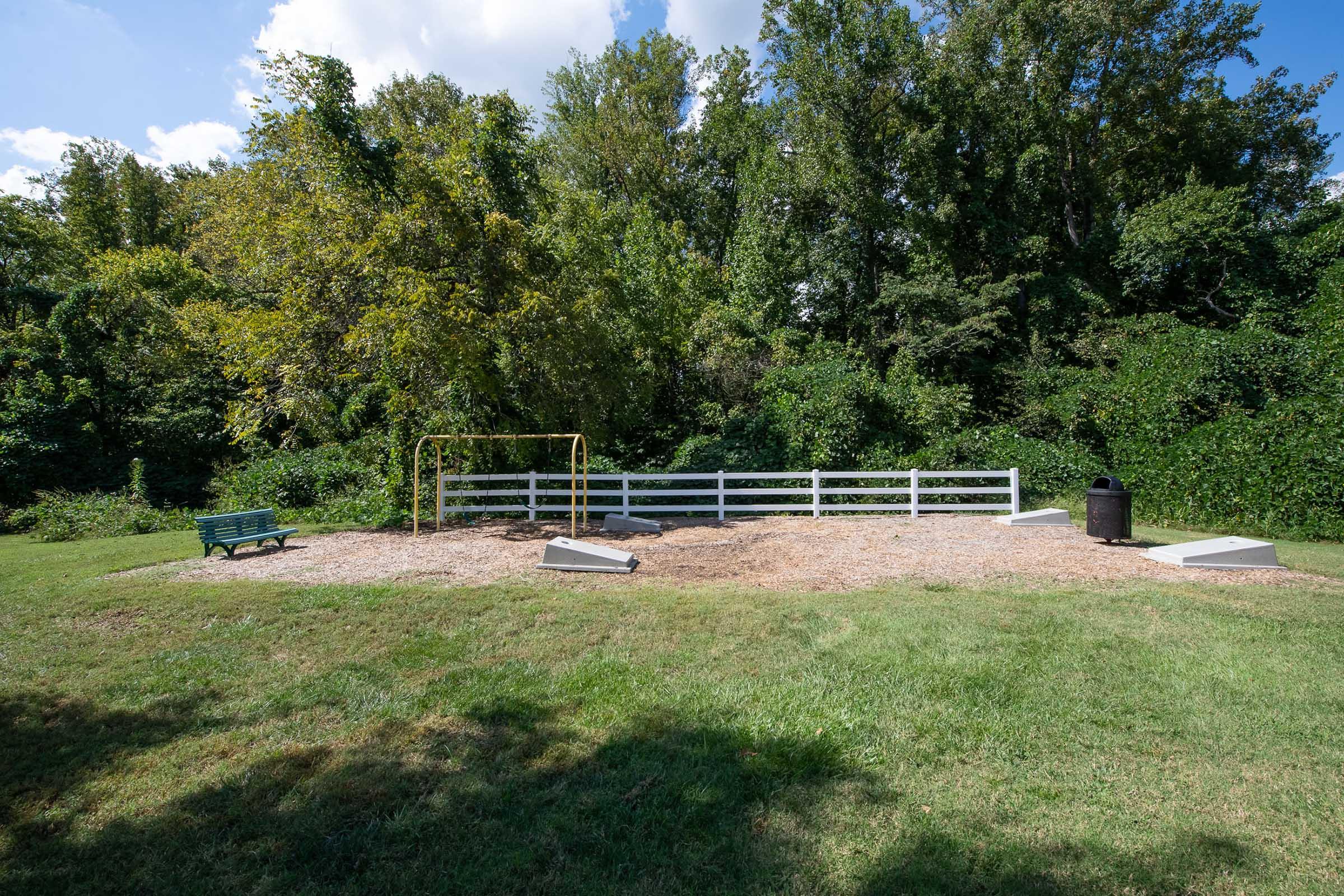 A small, empty playground area with a swing set and a bench near a wooded background. The ground is covered with mulch, and a trash can is visible. The setting is sunny with a few clouds.