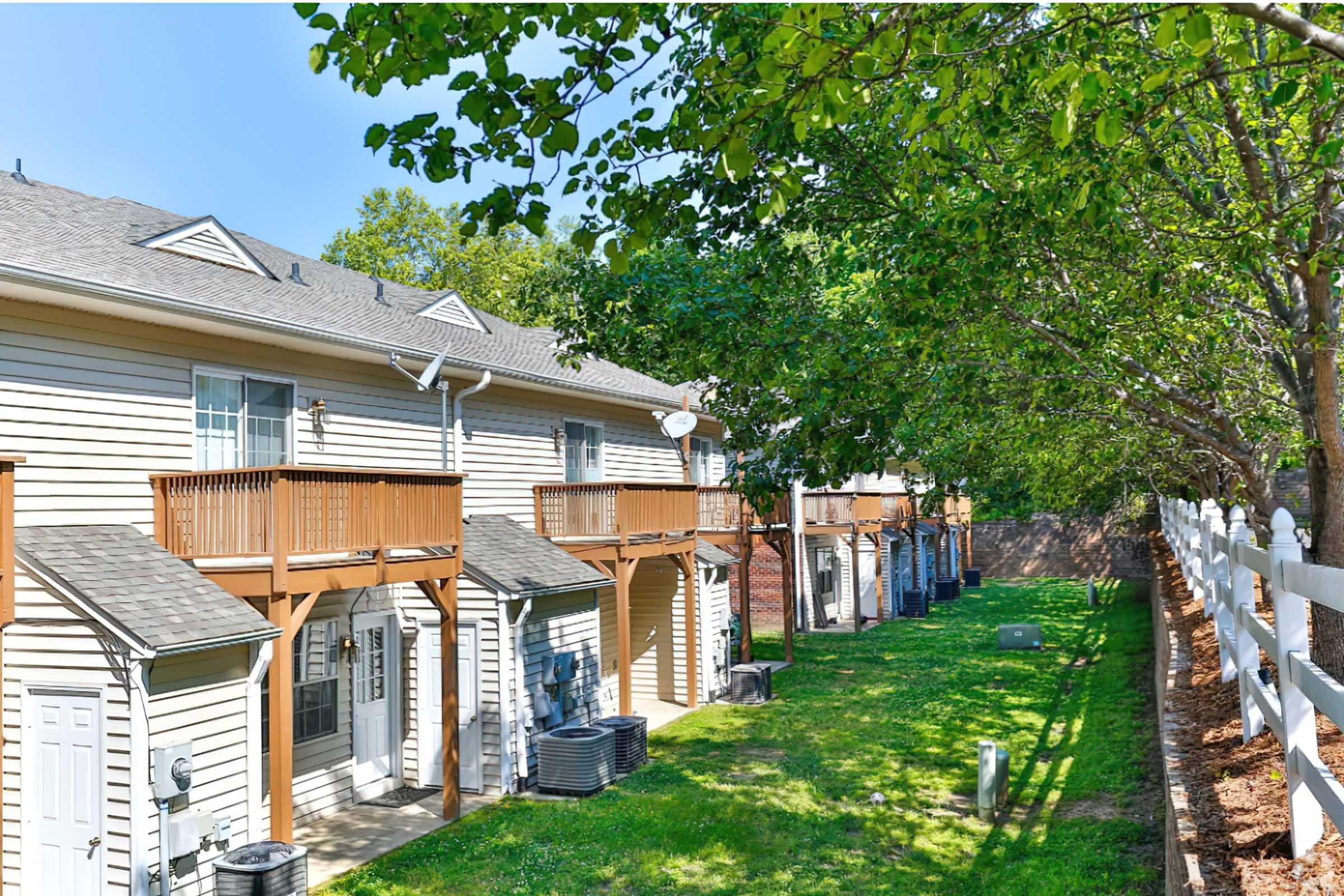 A view of a row of residential buildings, featuring balconies and air conditioning units, set in a well-maintained green area with trees and a white fence. The sky is clear and blue, indicating a sunny day.