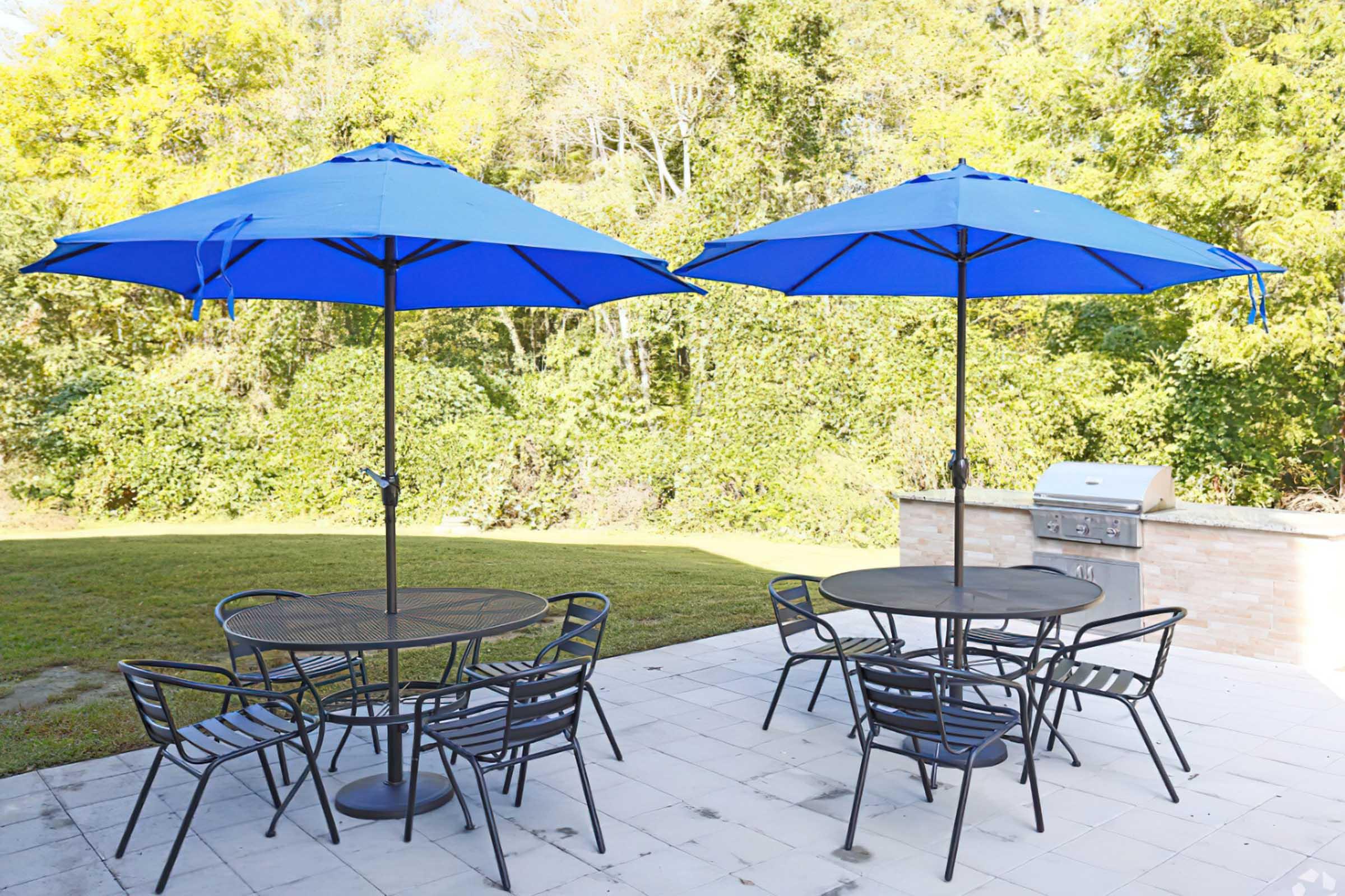 Outdoor patio scene featuring two round tables with black chairs, each shaded by a large blue umbrella. In the background, there's a grassy area and a stone barbecue grill, all set against a backdrop of lush green trees. The setting is bright and inviting, ideal for outdoor dining or gatherings.
