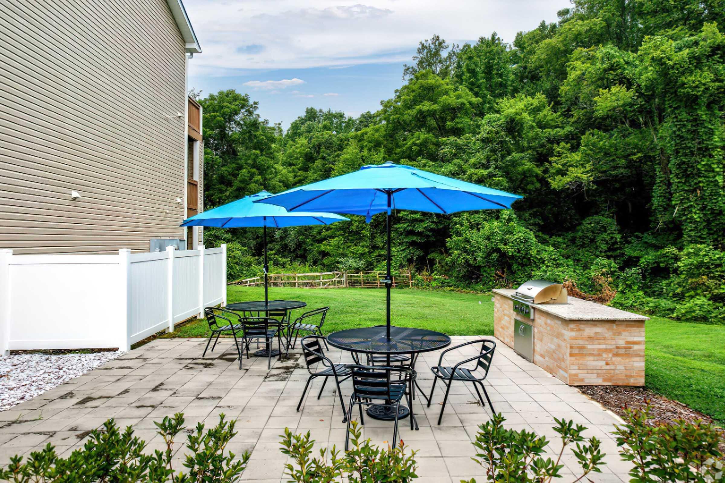 A backyard patio featuring two large blue umbrellas shading black metal tables and chairs. The area is surrounded by lush greenery, with a white fence in the background and a built-in grill on a stone surface nearby. The scene is bright and inviting, perfect for outdoor gatherings.
