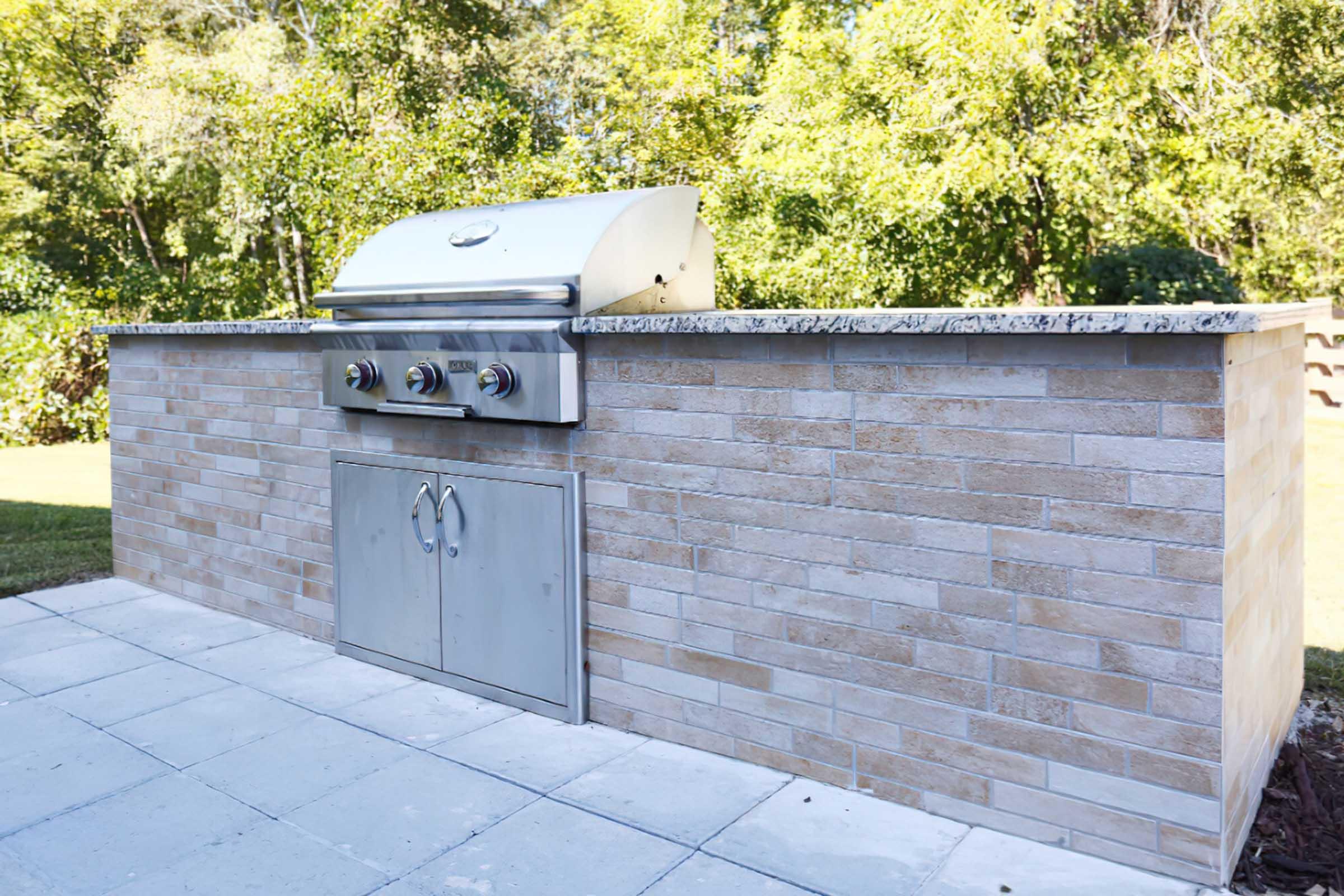 An outdoor kitchen setup featuring a stainless steel grill built into a stone countertop with a light-colored stone base. The grill has three knobs and a cabinet underneath for storage, surrounded by a grassy area and trees in the background.