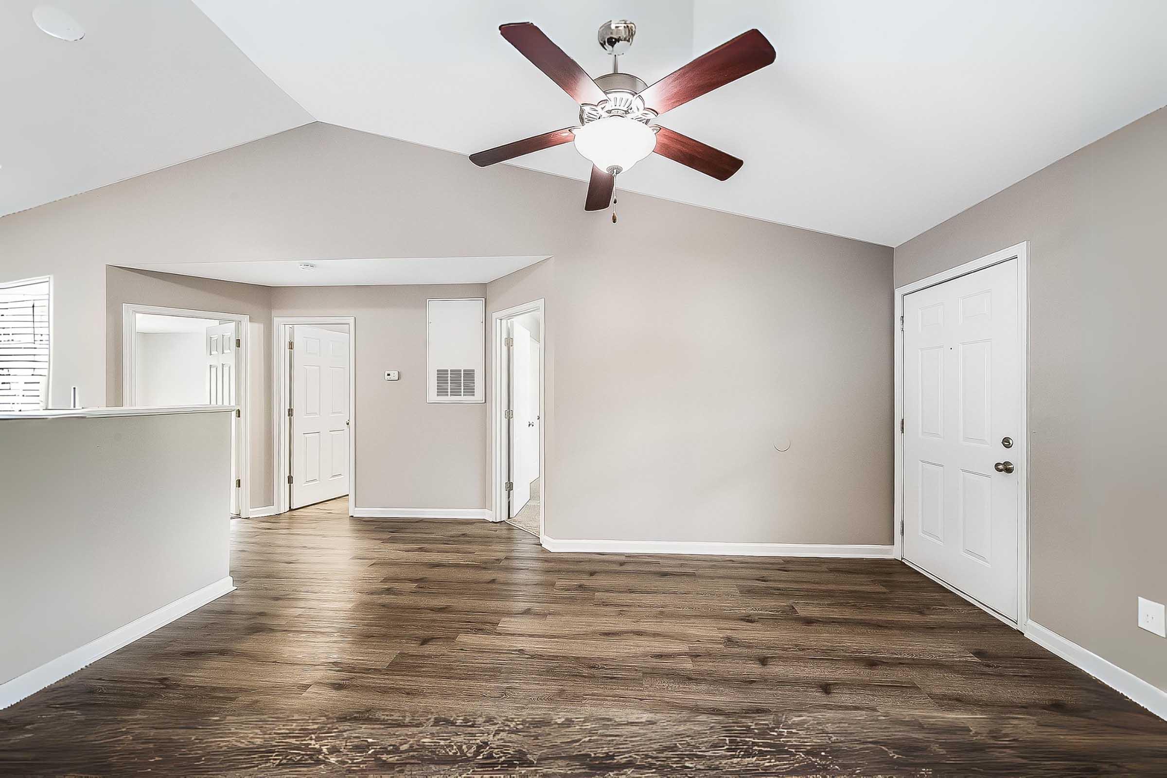 A spacious, well-lit living area with a ceiling fan, showcasing light-colored walls and dark wood flooring. Doorways lead to adjacent rooms, and a front door is visible on the right. The overall atmosphere is welcoming and modern.