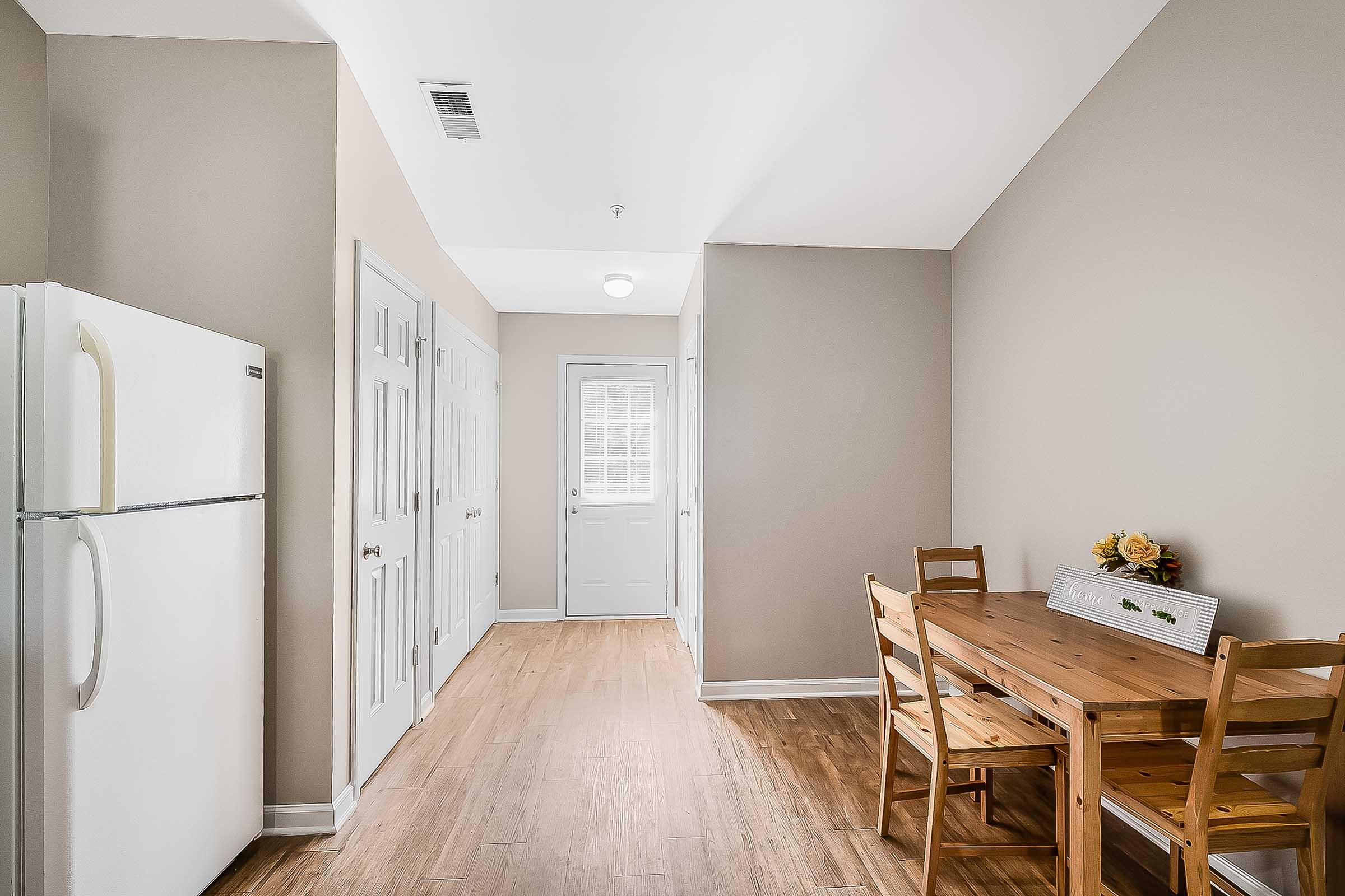 Bright and modern kitchen area featuring a white refrigerator, a wooden dining table with chairs, and a doorway leading outside. Light-colored walls and wooden flooring create a warm and inviting atmosphere. Additional cabinets line one wall for storage.