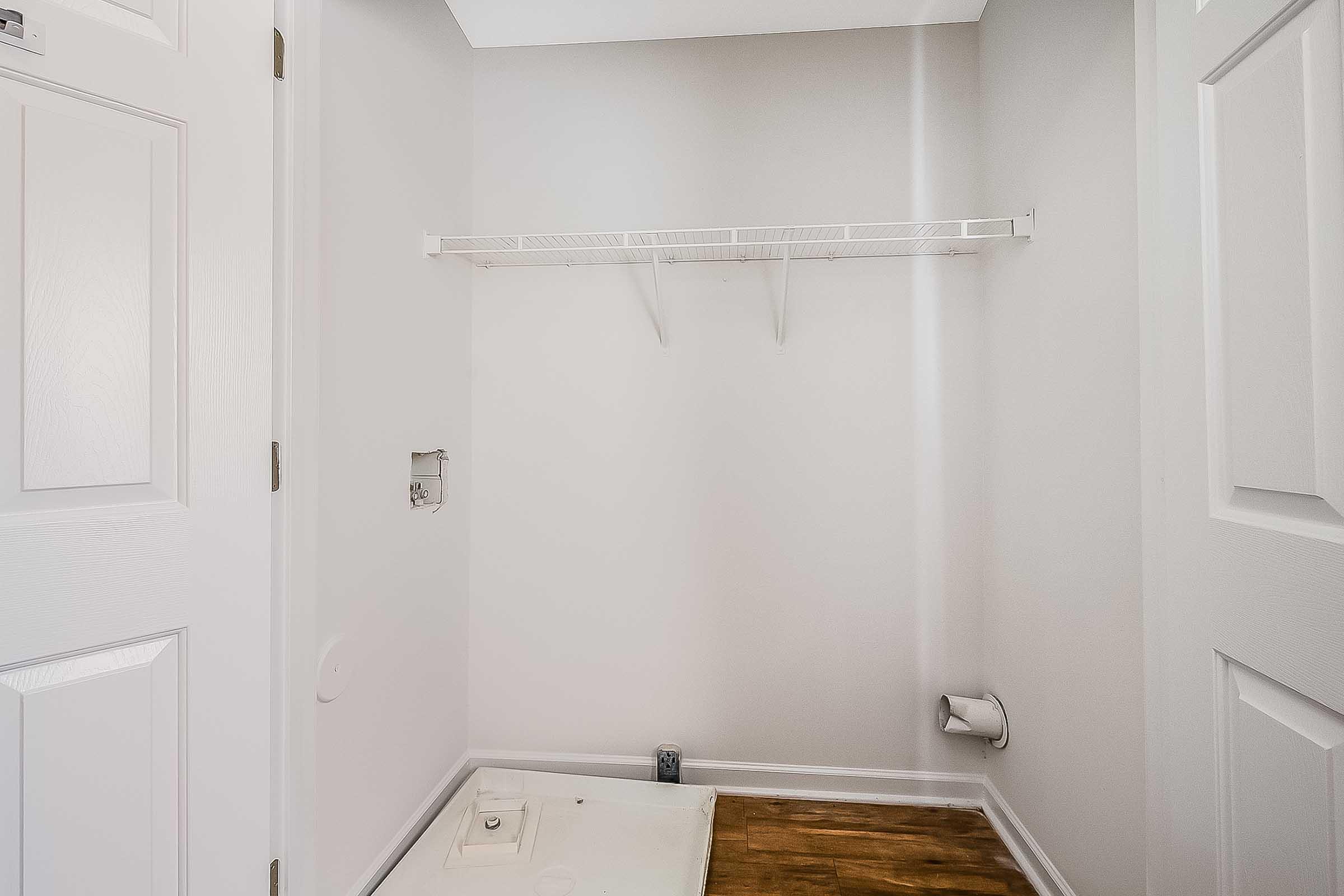 A vacant laundry room with light gray walls, featuring a white wire shelf against the wall and a small empty space for a washing machine. The floor is wooden, and there's a plumbing outlet visible on the right side. The door to the room is closed.
