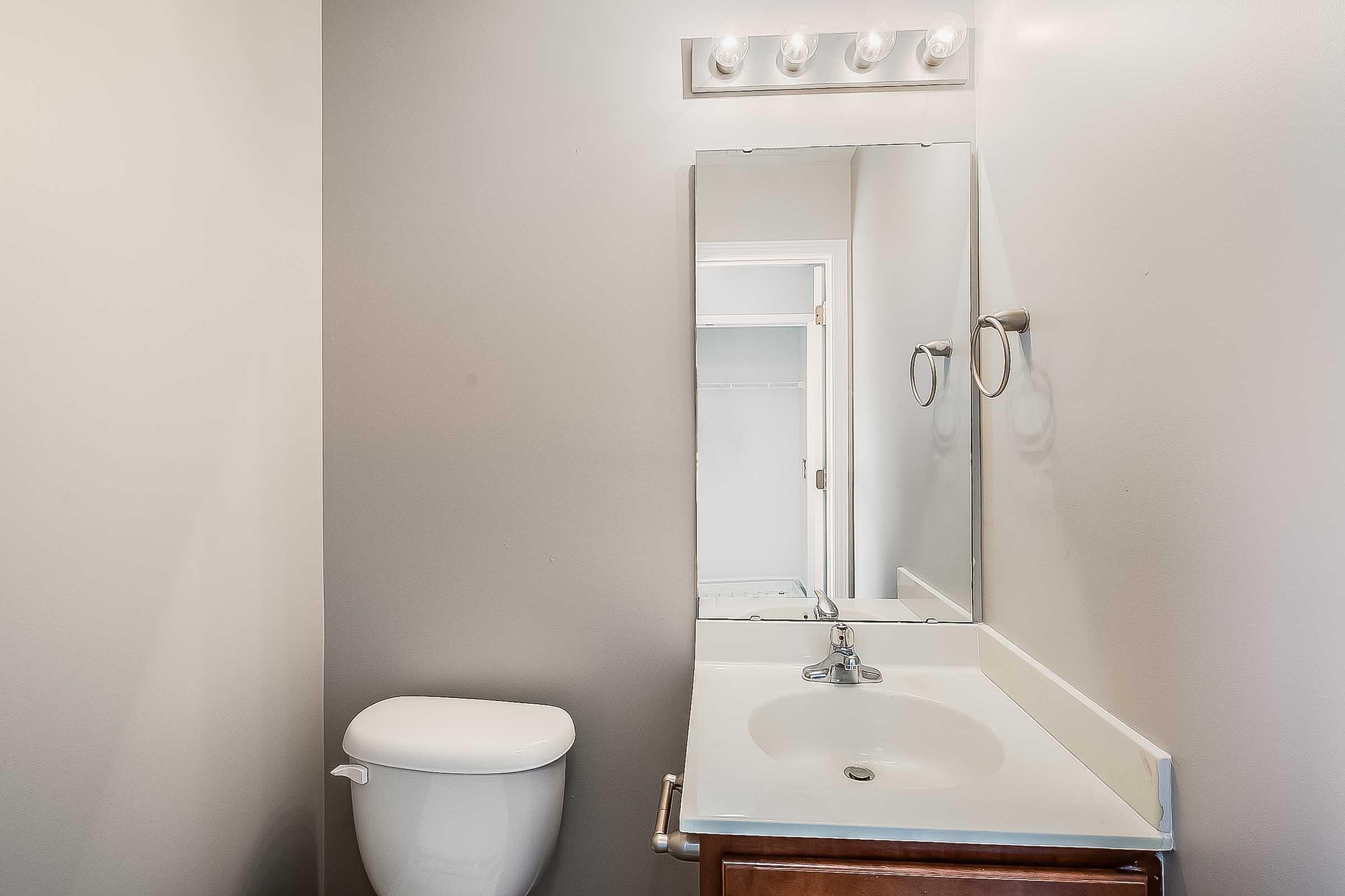 A clean, neutral-toned bathroom featuring a white toilet and a modern sink with a countertop. Above the sink, there is a large mirror and a light fixture with multiple bulbs. The wall color is light gray, creating a calming atmosphere. A glimpse of a door to a closet is visible in the background.