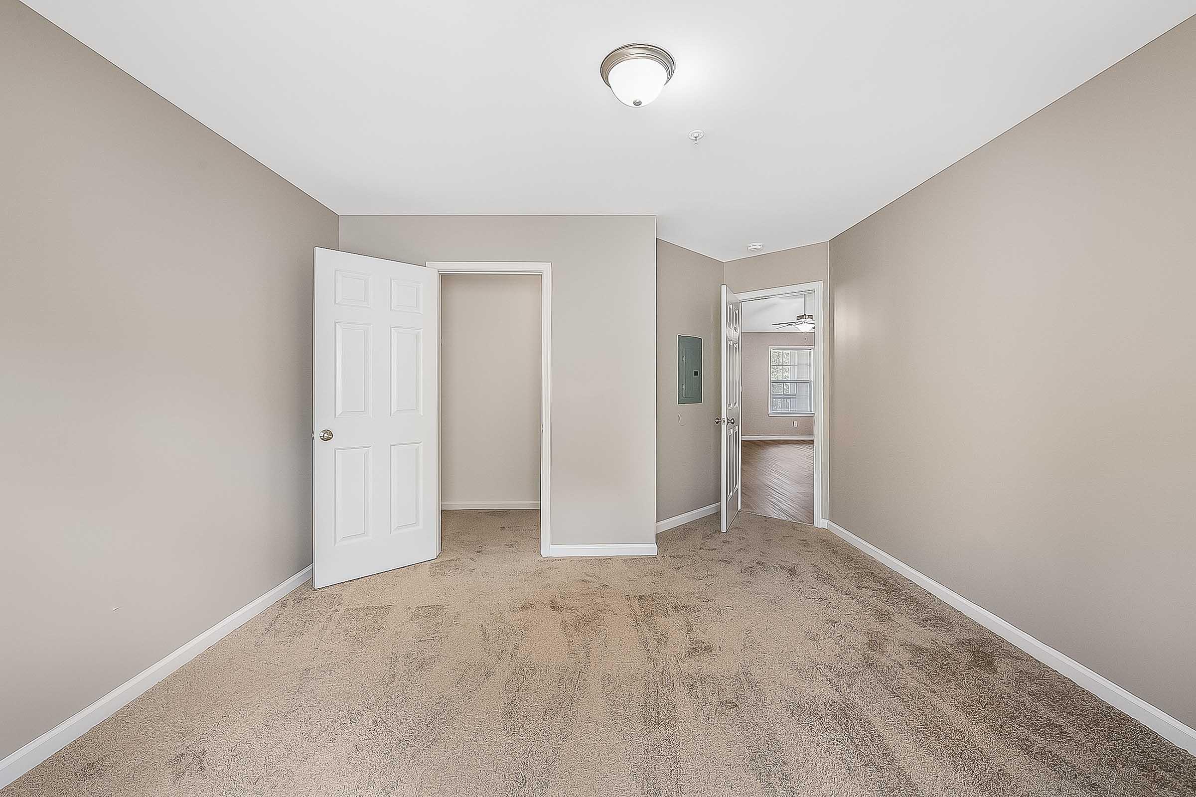 Interior view of a room with beige walls and carpet. There is a door on the left leading to a small closet, and another door on the right that opens into a hallway or another room. A light fixture is visible on the ceiling, and natural light is coming in from the door at the end of the hallway.