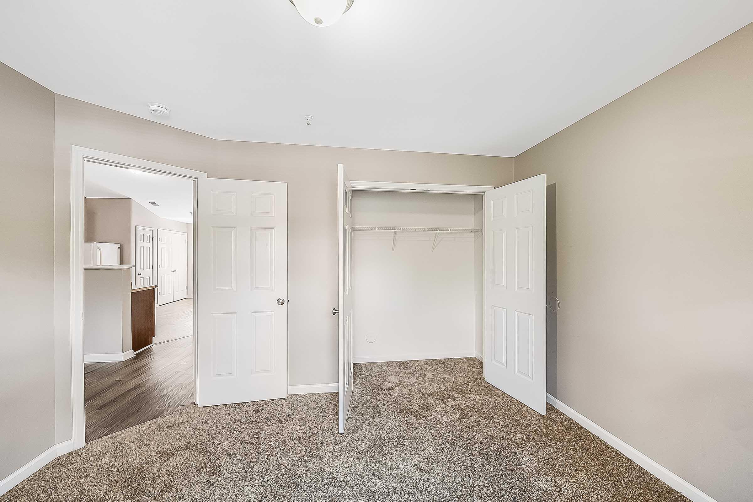 Empty room with beige walls and carpeted floor. Two white closet doors are open, revealing an uncluttered closet with a hanging rod. The room has a ceiling light fixture and a doorway leading to another area, with light coming from the hall. Overall, it's a clean and spacious environment.