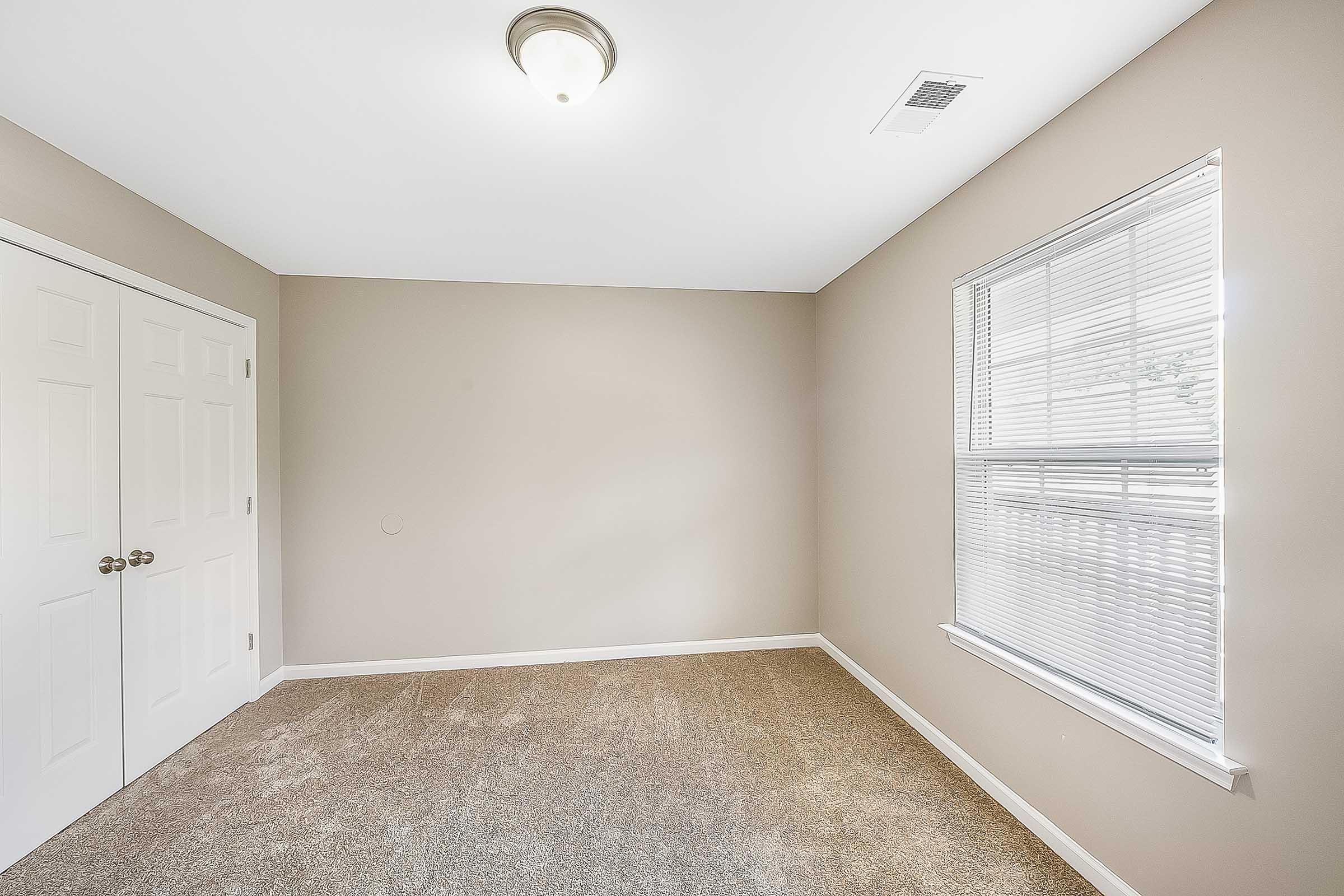 Empty room with light beige walls and carpet, featuring a window with blinds on one side and a double-door closet on the other. A ceiling light is installed, providing overhead illumination. The room has a bright and airy feel due to natural light coming through the window.