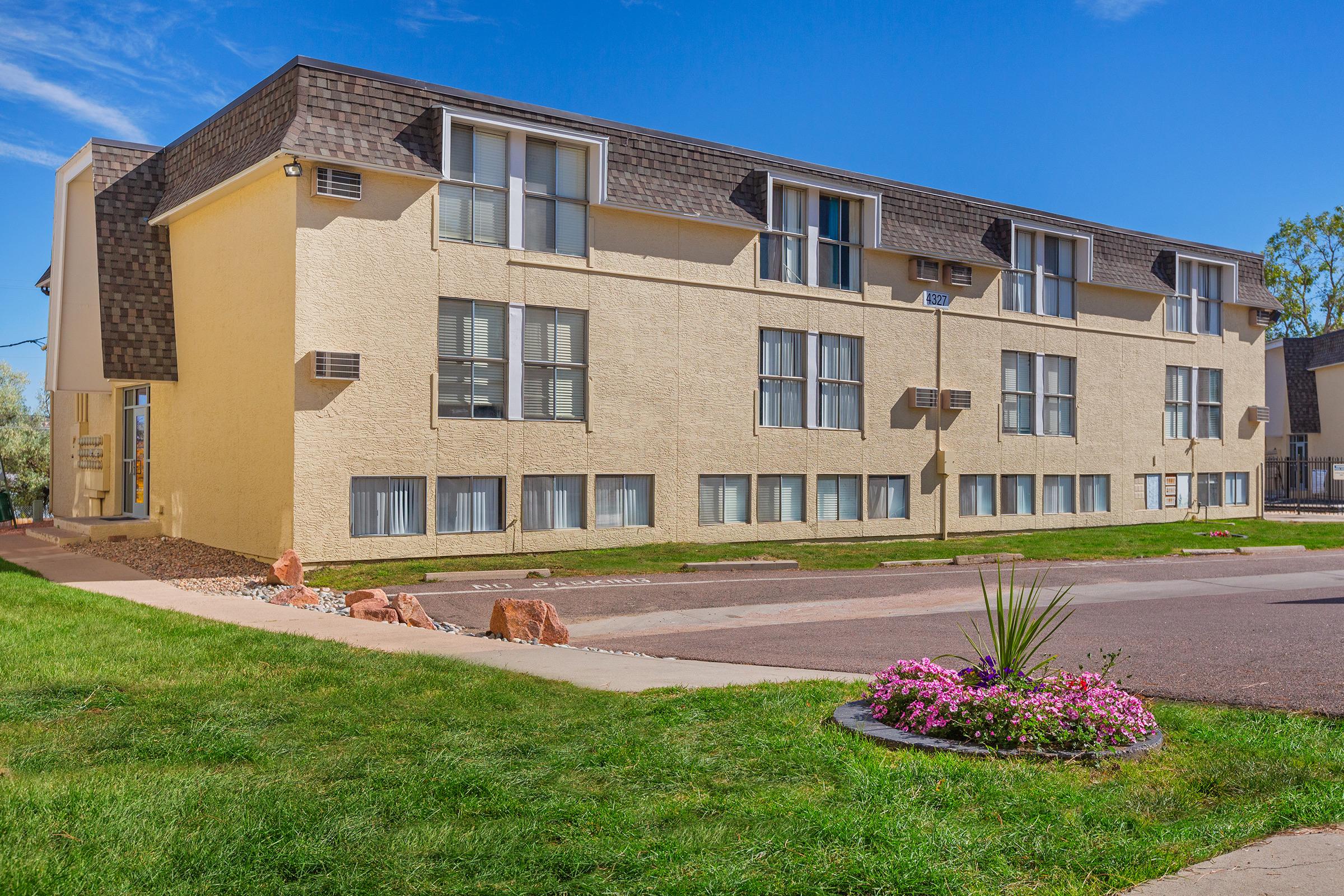 A two-story beige apartment building with multiple windows on the facade. The building features a sloped roof and is surrounded by well-maintained grass and a flower bed with pink flowers and greenery in front. The sky is clear and blue, adding to the pleasant appearance of the surroundings.