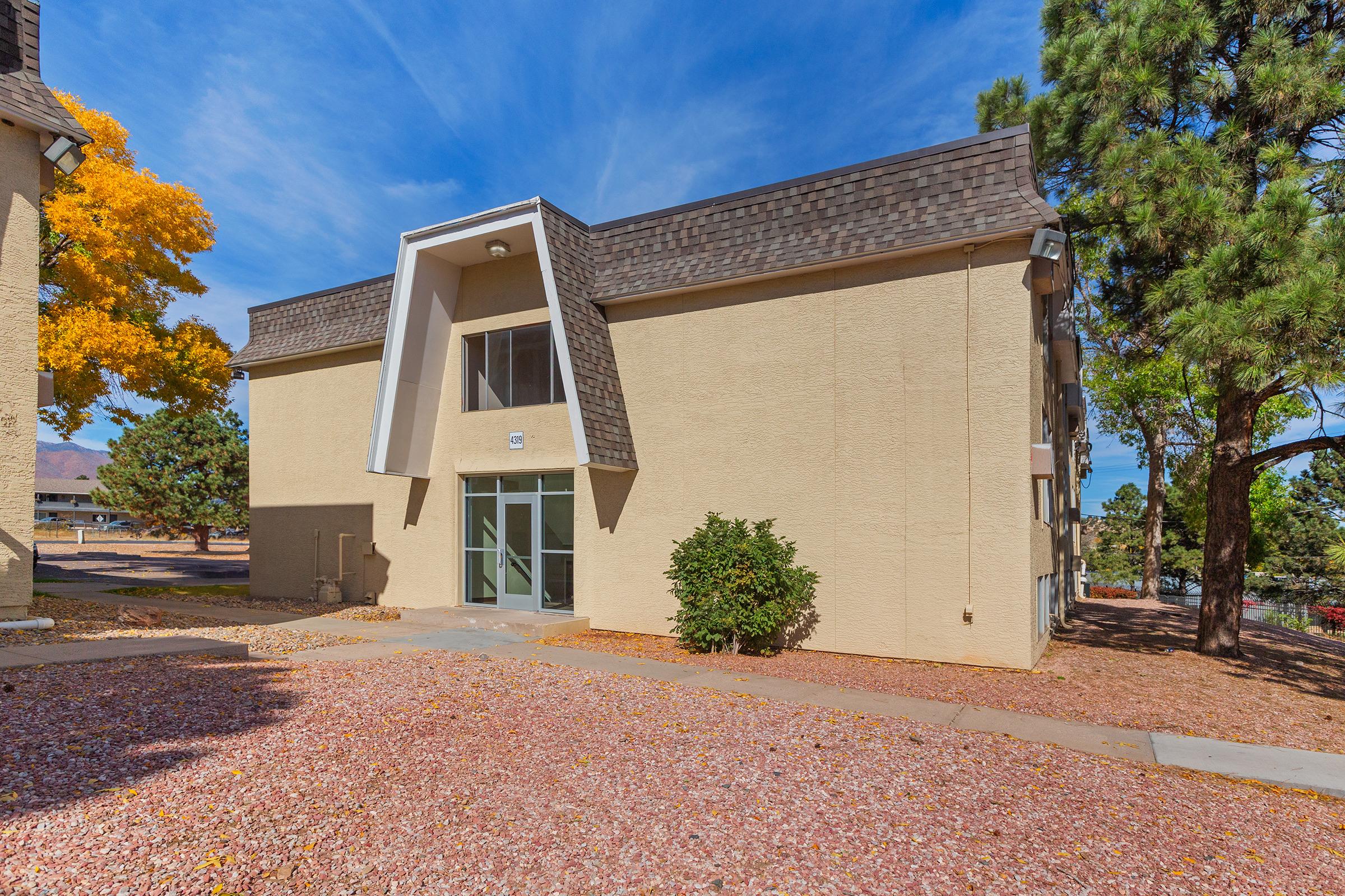 A modern two-story building with a unique triangular entrance, surrounded by a landscaped area featuring gravel and trees. The building has a beige exterior and is set against a blue sky with scattered clouds and autumn foliage on nearby trees.