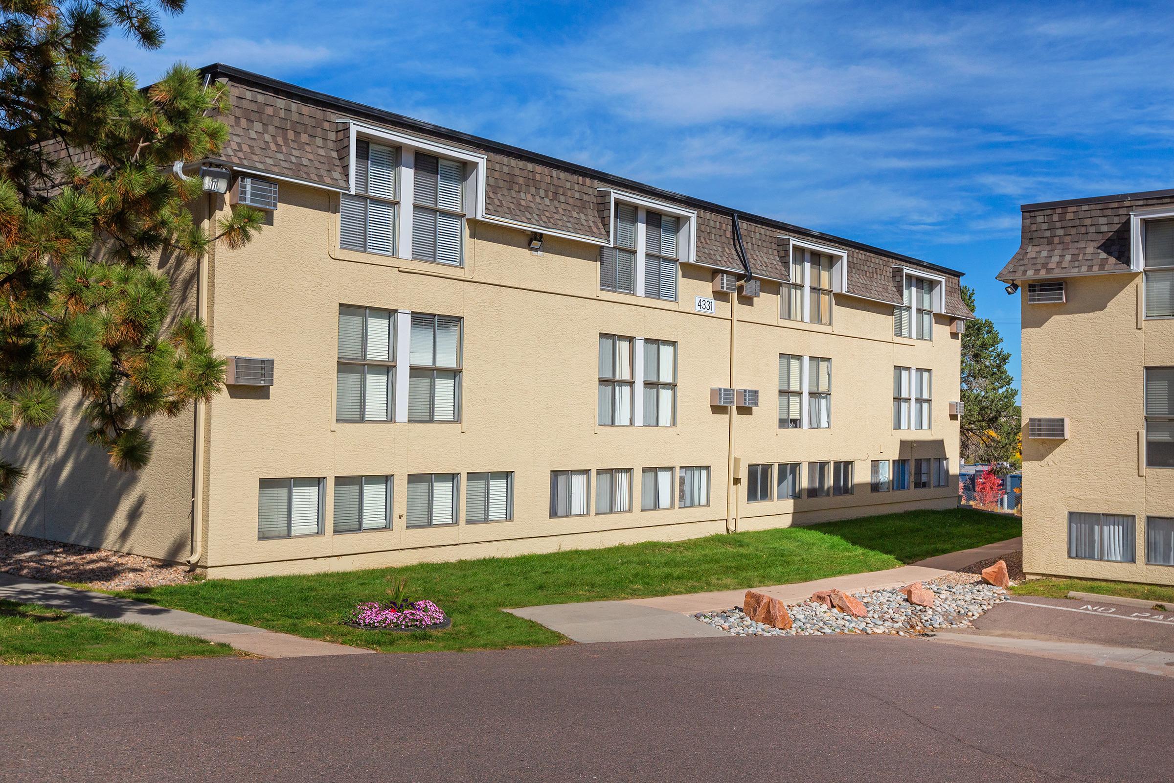 A multi-story residential building with light-colored exterior walls. The building features several windows with white shutters. A landscaped area includes grass and flower beds, while a concrete path leads up to the entrance. The sky is clear and blue, creating a bright atmosphere.