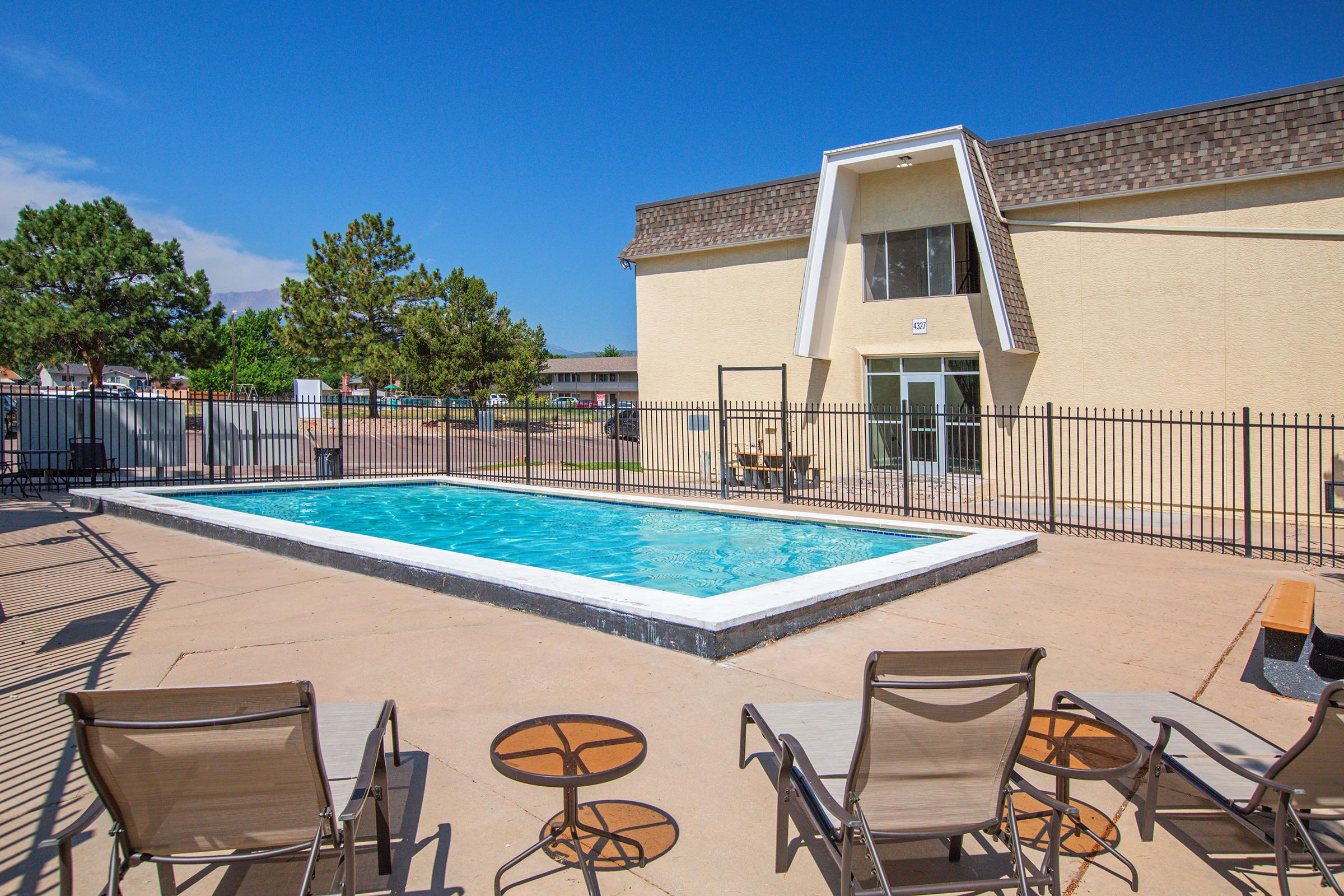 A clear blue swimming pool surrounded by a fenced area, with lounge chairs and small side tables on a concrete deck. A beige building with large windows is visible in the background, under a bright blue sky with a few clouds. Trees and additional buildings are seen in the distance.