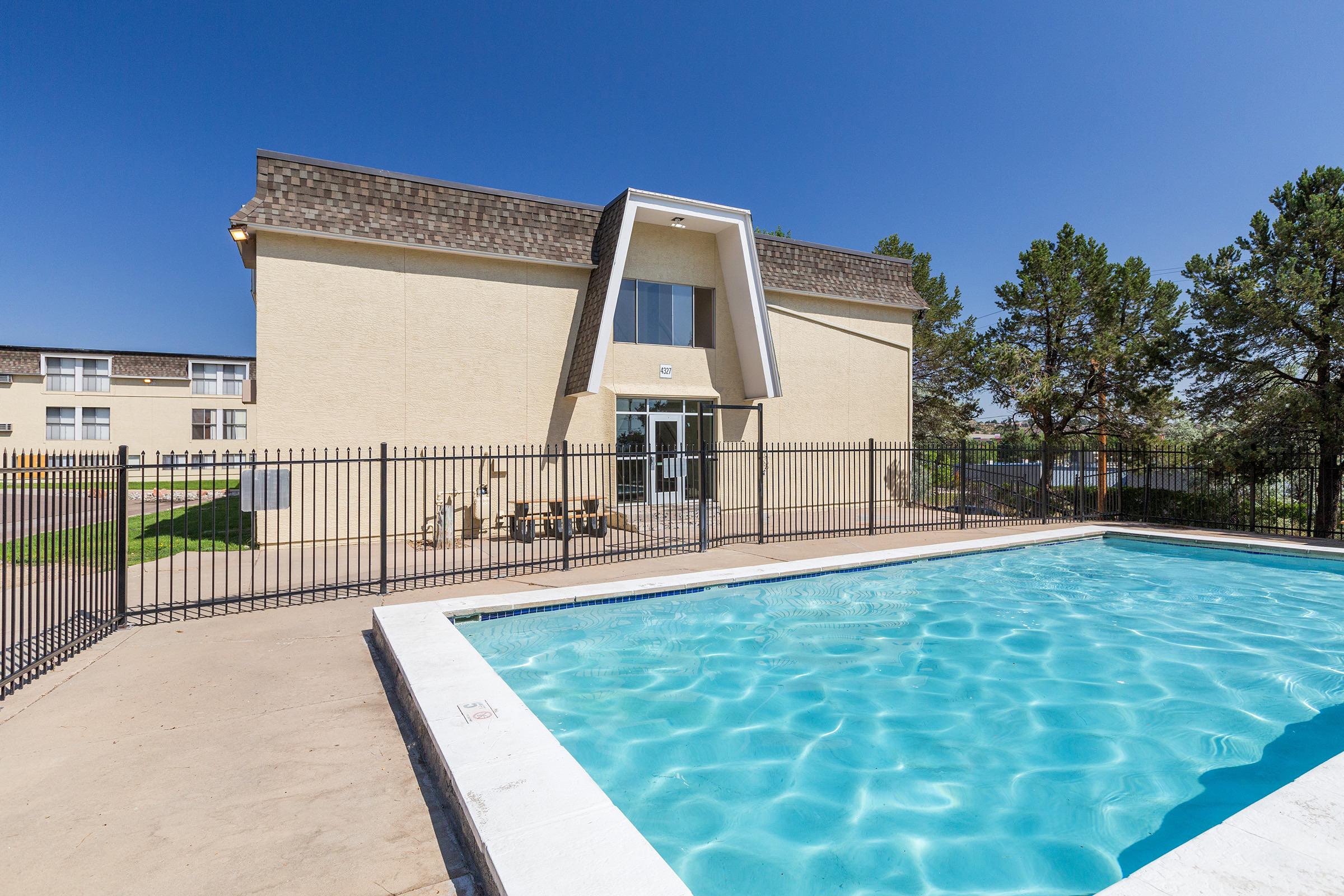 A clear blue swimming pool surrounded by a fence, with a light-colored building in the background. The building features large windows and a sloped roof. The scene is set under a bright blue sky, with a few trees visible nearby, suggesting a sunny outdoor area.