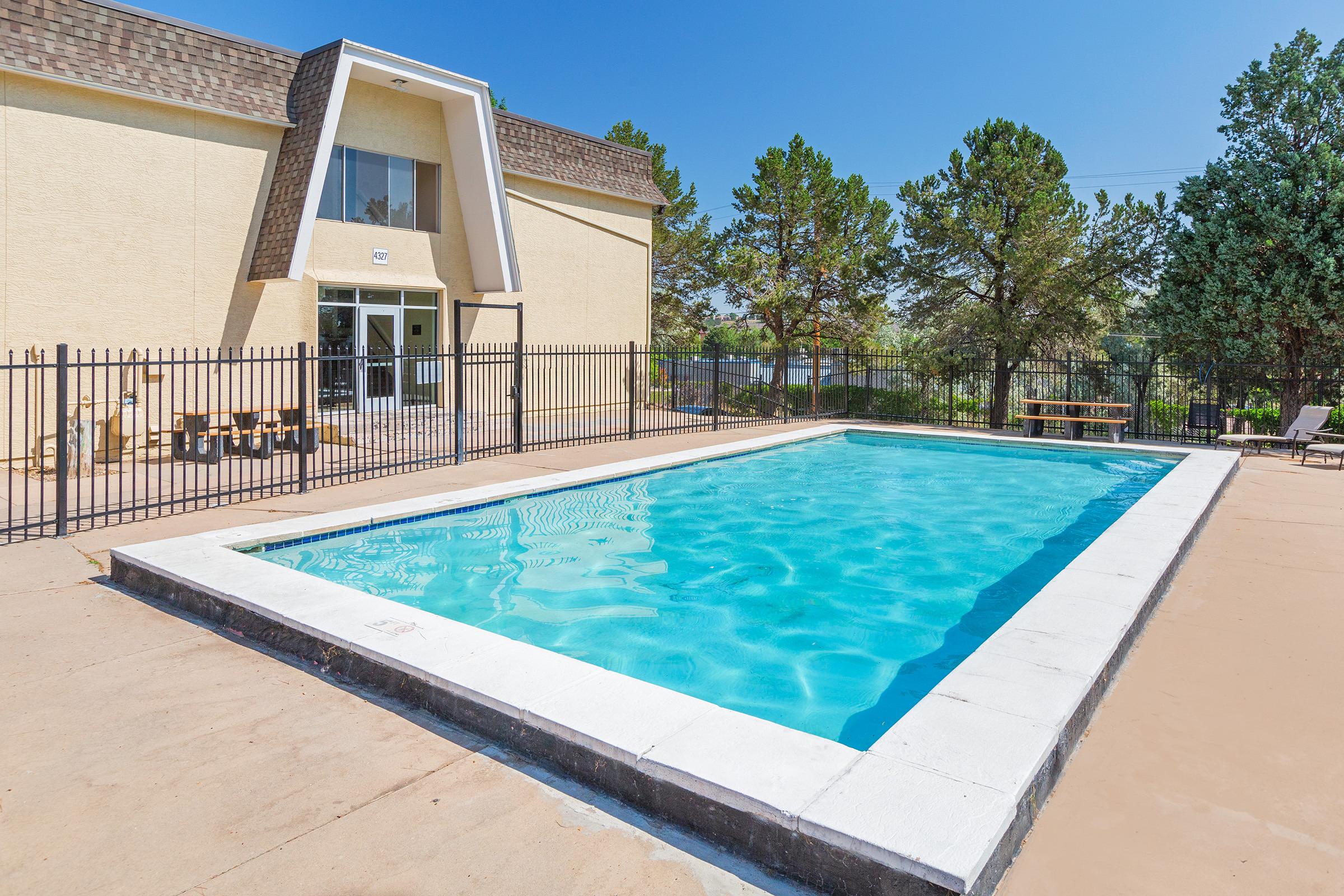 A clean and inviting outdoor swimming pool surrounded by a concrete patio. The pool features clear blue water and is enclosed by a black fence. In the background, there is a two-story building with large windows, trees providing shade, and a few lounge chairs set up for relaxation.