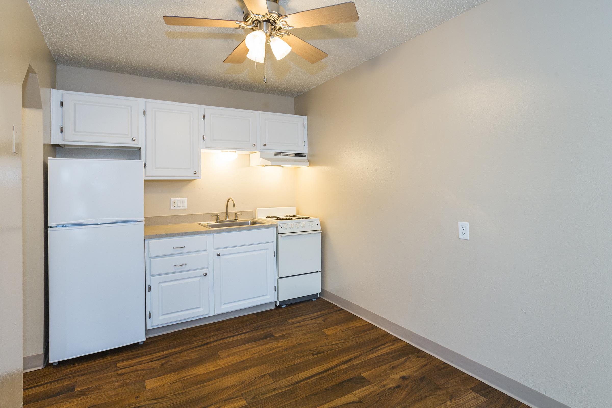 A small kitchen featuring white cabinetry, a stainless steel sink, a refrigerator, and a stove. There's a ceiling fan above, and the floor is made of wood laminate. The walls are painted a light beige color, creating a cozy atmosphere.