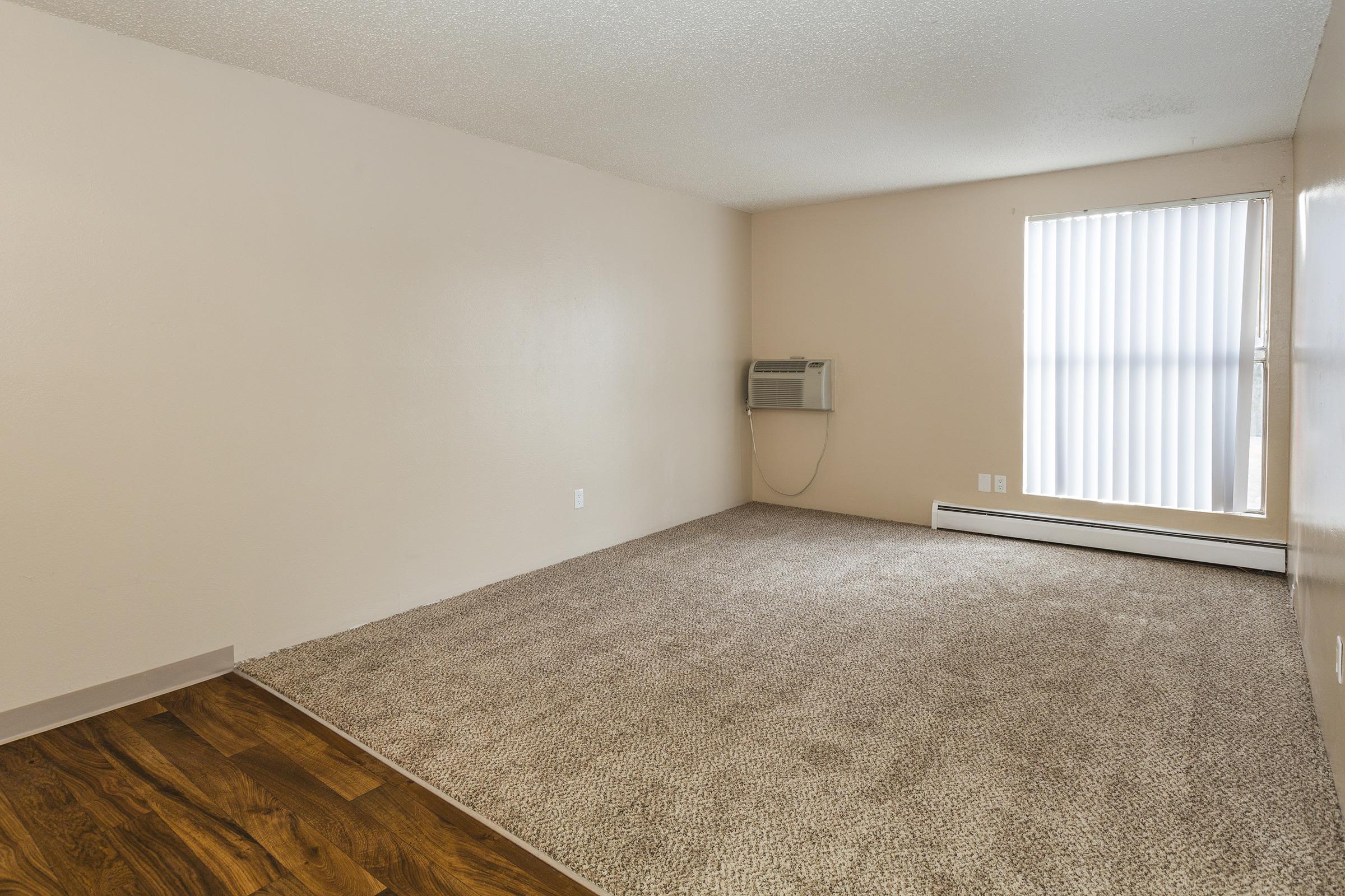 A spacious, empty room with light beige walls and a soft carpeted floor. There is an air conditioning unit on the wall and a window with white vertical blinds, allowing natural light to enter. The room is simple, with no furniture, highlighting the open space.
