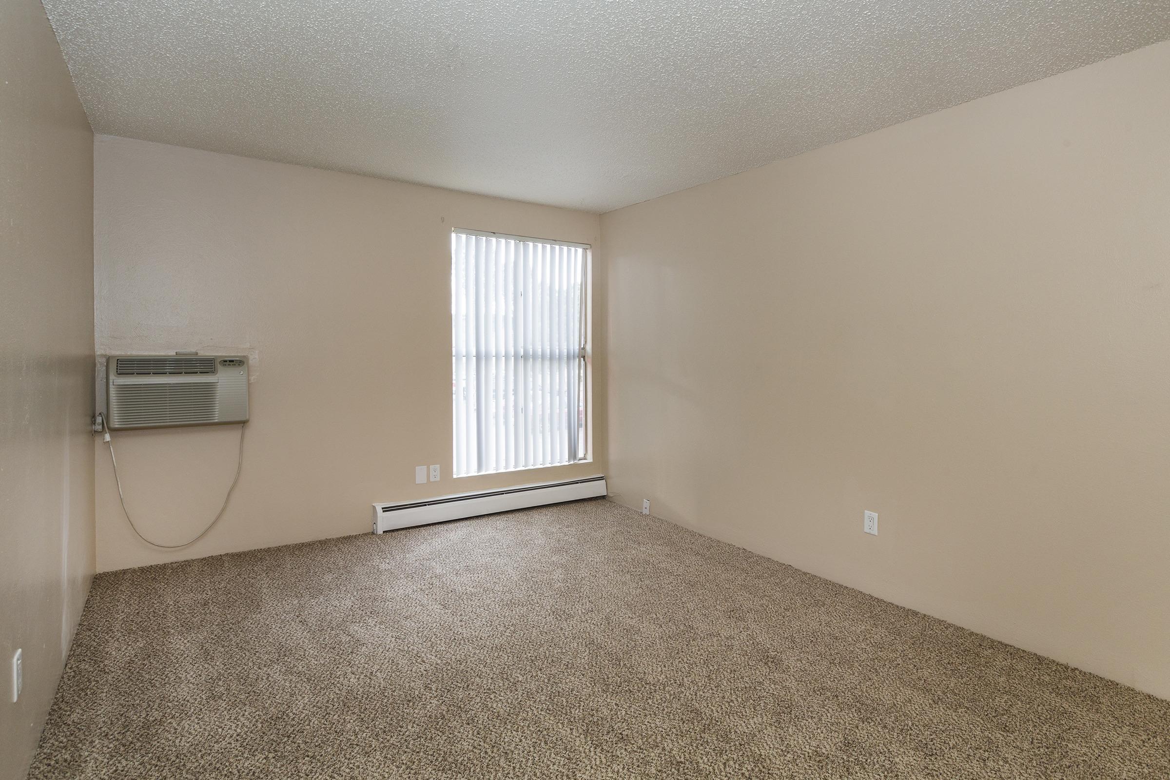 A vacant room featuring beige walls and carpeted flooring. There is a single window with vertical blinds allowing natural light, and an air conditioning unit mounted on the wall. The space appears clean and ready for furnishing.