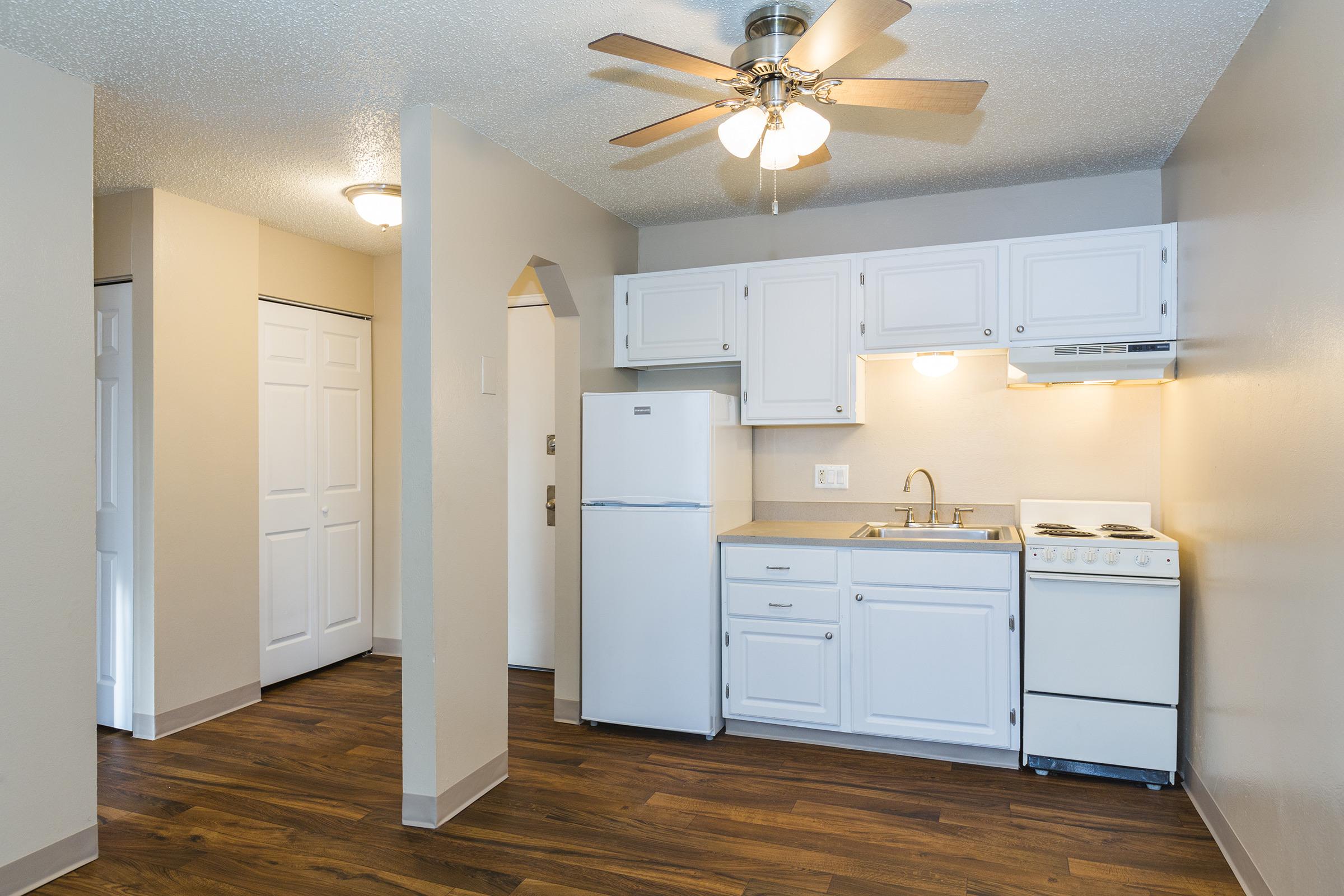 A small kitchen area featuring white cabinetry, a refrigerator, a stove, and a sink. The space has wood-look flooring and a ceiling fan. Nearby, there's an open doorway leading to another room with a closet, and the walls are painted in neutral tones. Natural light illuminates the area.