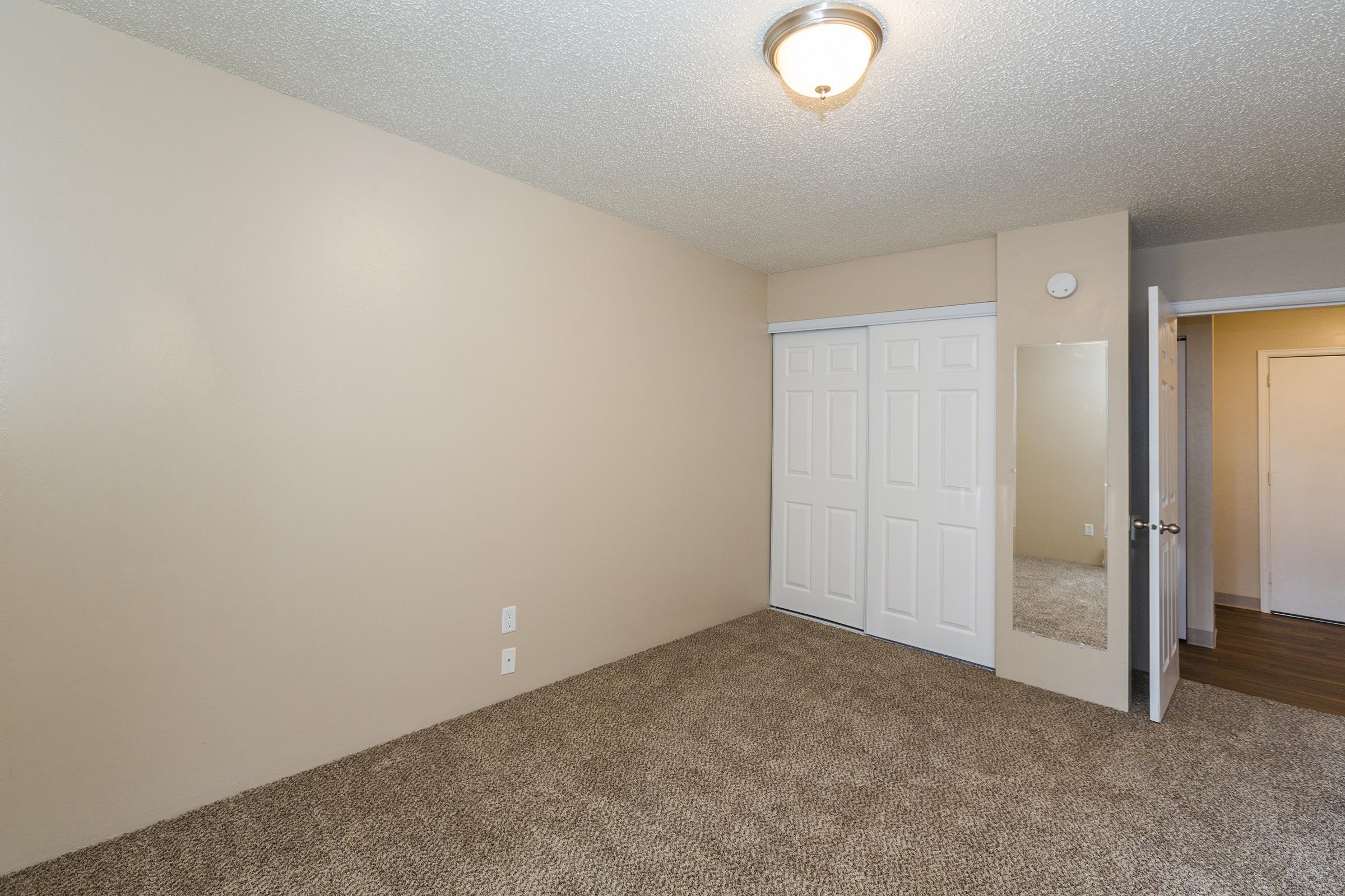 A spacious, empty room with beige walls and carpeted flooring. The room features a ceiling light fixture, a mirrored closet door, and a second door leading to another area. The overall atmosphere is bright and clean, suitable for various uses.