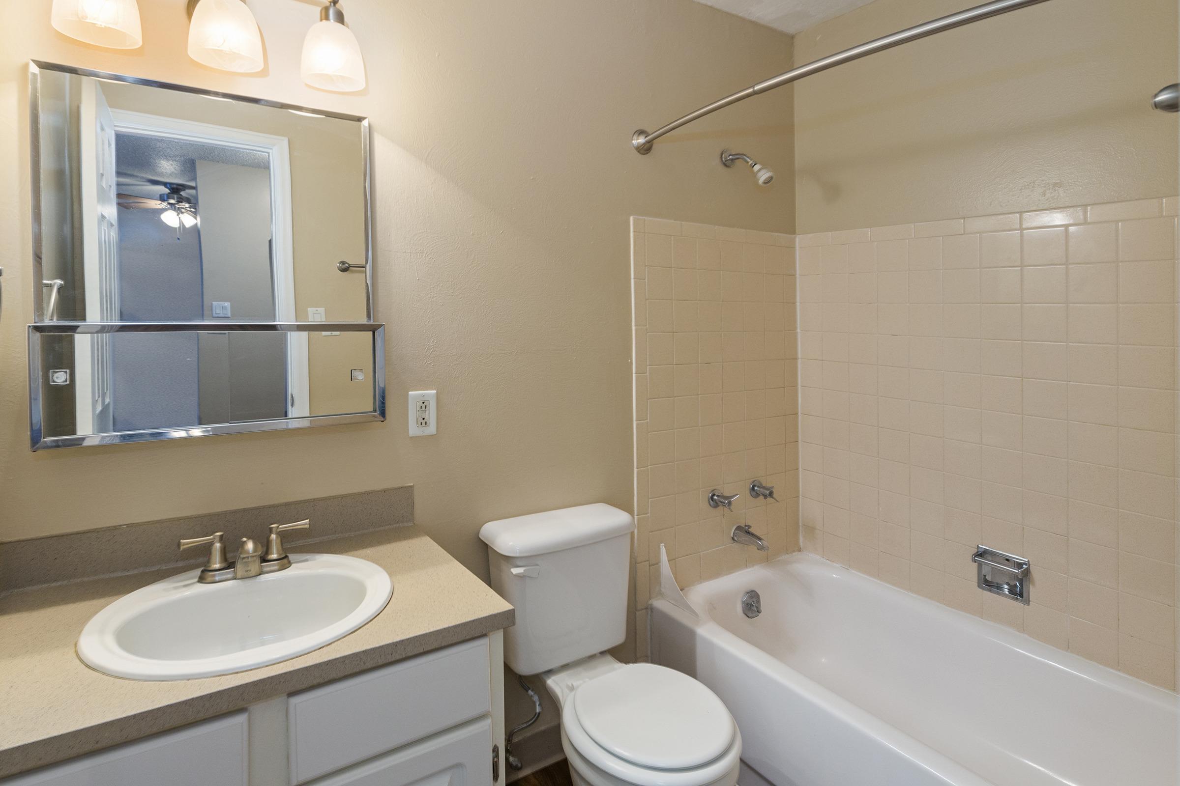 A clean and neutral bathroom featuring a bathtub with a shower, a toilet, and a sink with a mirror above it. The walls are painted a light beige color, and the tiles in the shower area are arranged in a grid pattern. Soft lighting is provided by three overhead fixtures.