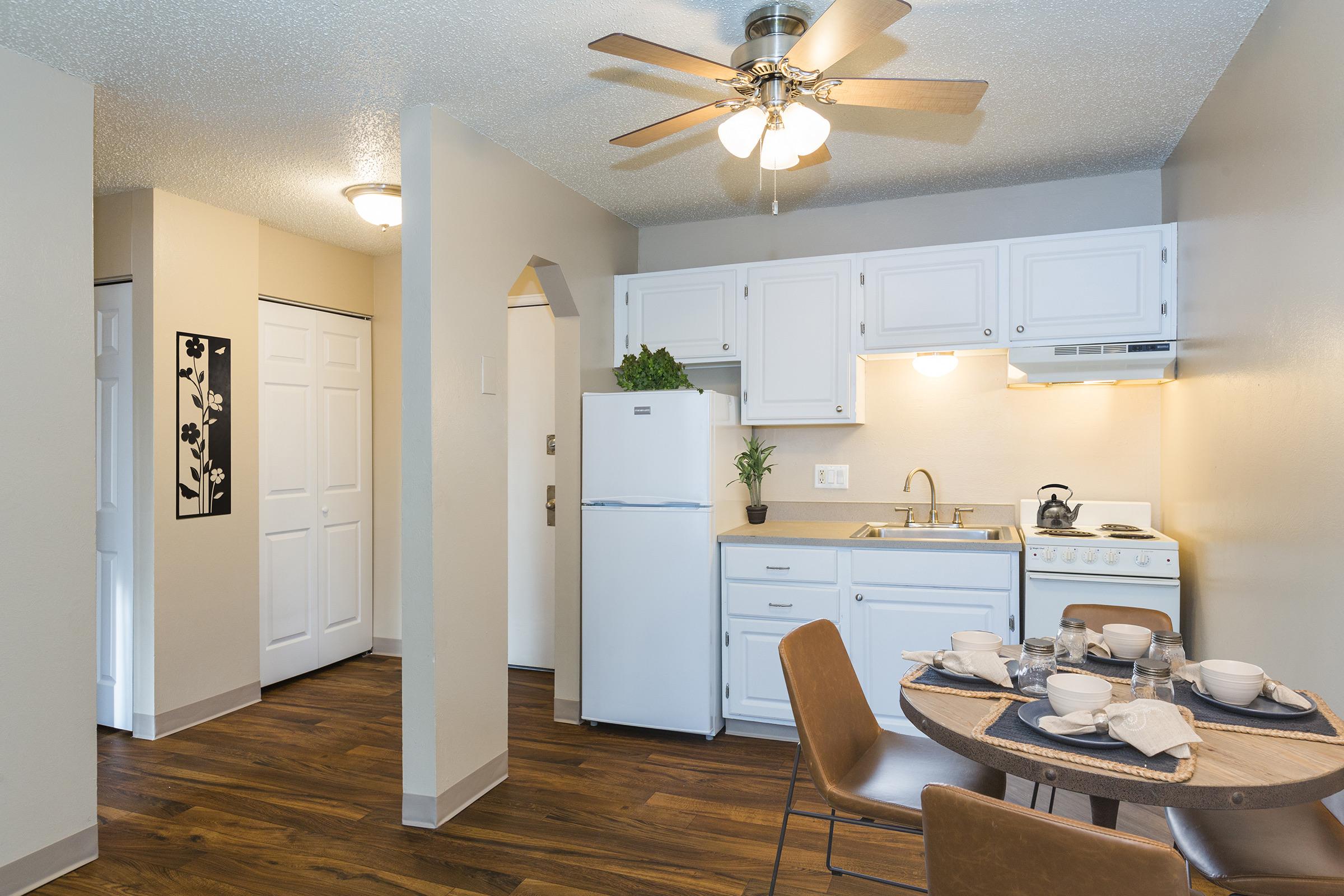 Modern kitchen featuring white cabinets and appliances, including a refrigerator and stove. A dining table set for four is in the foreground, with stylish dinnerware. The space has warm wooden flooring and a ceiling fan, with a door leading to another room in the background.