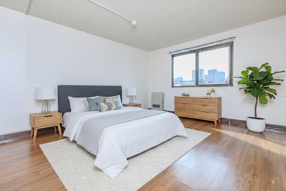 A modern bedroom featuring a neatly made bed with gray and white bedding, two bedside lamps on wooden nightstands, a stylish wooden dresser, and a small potted plant. Large windows allow natural light and offer a view of the city skyline. The room has light-colored walls and hardwood flooring.