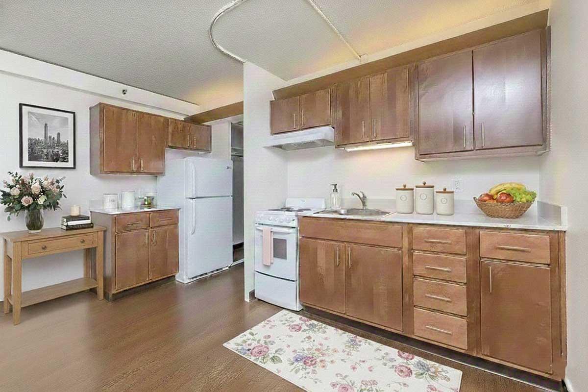 A modern kitchen featuring wooden cabinets, a white stove, and a refrigerator. There is a small table with flowers, a countertop with decorative jars, and a bowl of fruit. The floor is covered with a floral rug, and the overall design is bright and inviting.