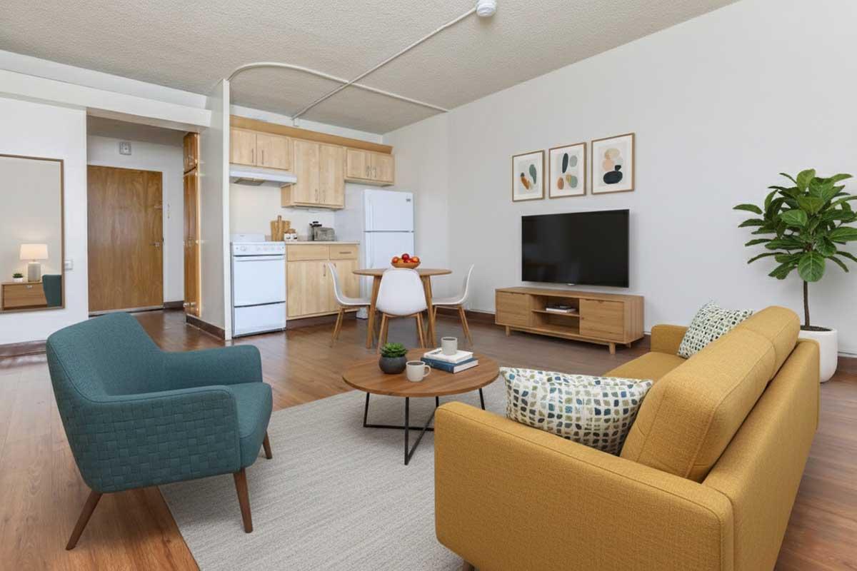 A modern living room featuring a yellow sofa and a teal armchair, a round coffee table with a plant and decorative items, and a TV stand. In the background, there is a kitchenette with wooden cabinetry, a white refrigerator, and dining chairs. The walls are adorned with framed artwork and a potted plant adds greenery.