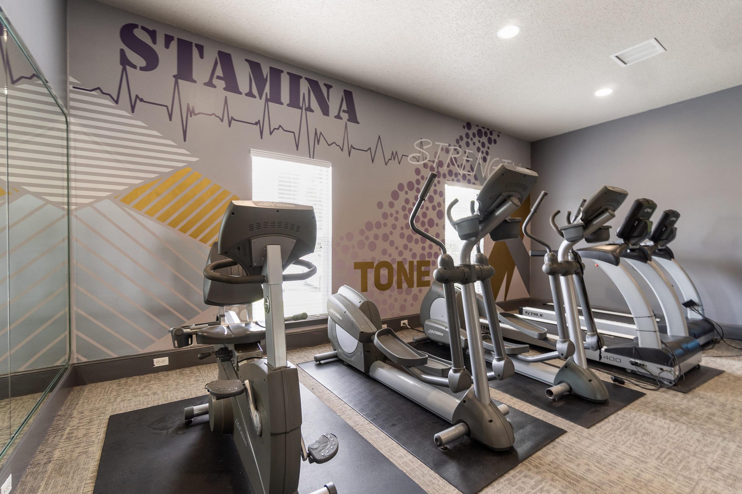 A modern fitness gym featuring several cardio machines including ellipticals and stationary bikes, set against a motivational wall with the words "STAMINA" and "TONE." The space is well-lit, with large mirrors and a clean, inviting atmosphere.
