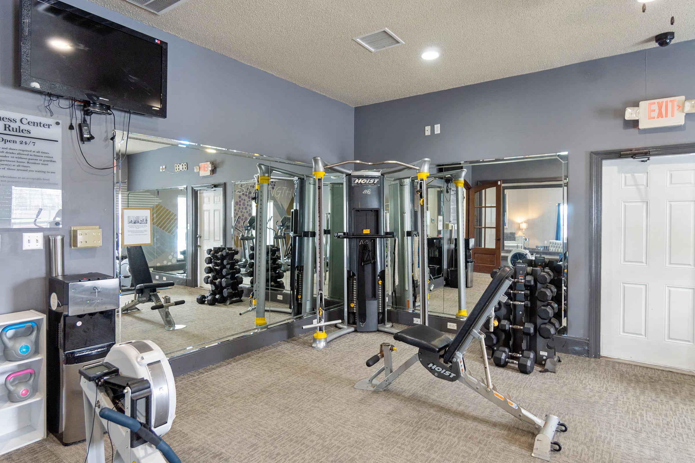 A small gym area featuring a weight bench, free weights, and a multi-station exercise machine. The walls are mirrored to create a spacious feel, and there is a wall-mounted TV. The flooring is carpeted, and the overall decor is neutral with a hint of modern style. A door leads to another room in the background.