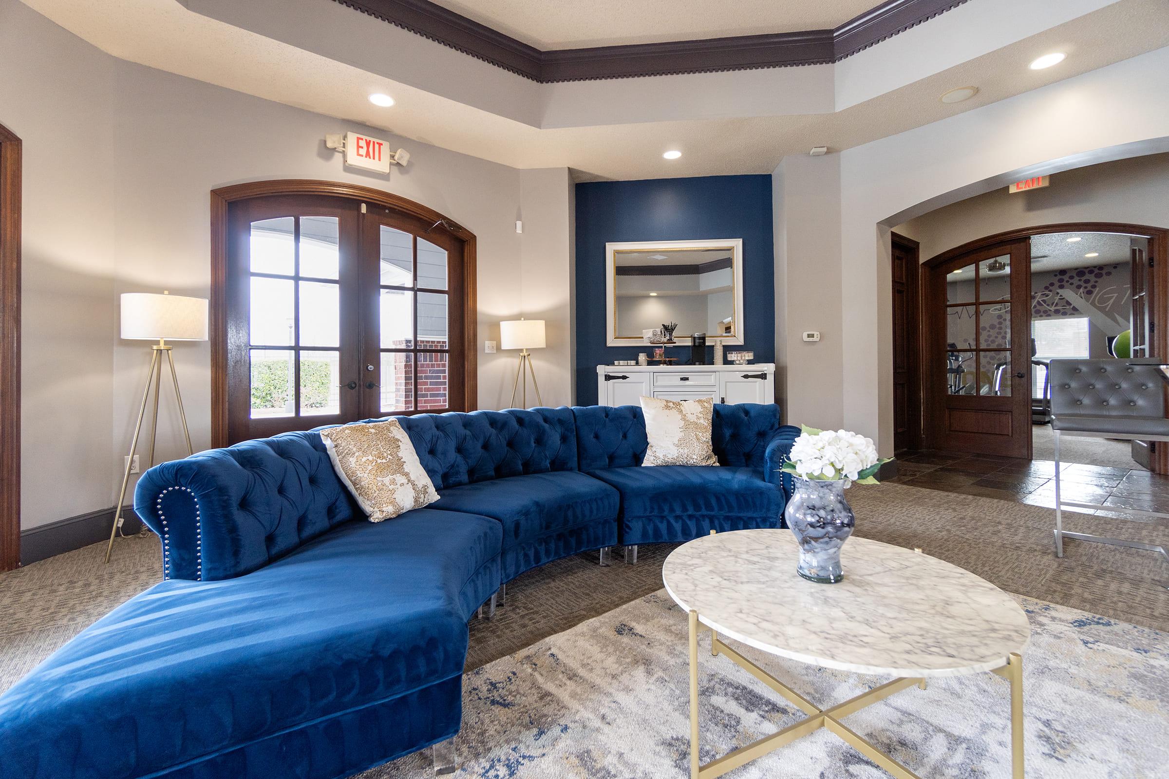 A stylish lounge area featuring a curved blue velvet sofa with decorative pillows, a marble coffee table, and fresh flowers in a vase. The space is well-lit with large windows, showcasing a modern, elegant design with blue and white accents. Decor includes a mirror and tasteful artwork on the walls.