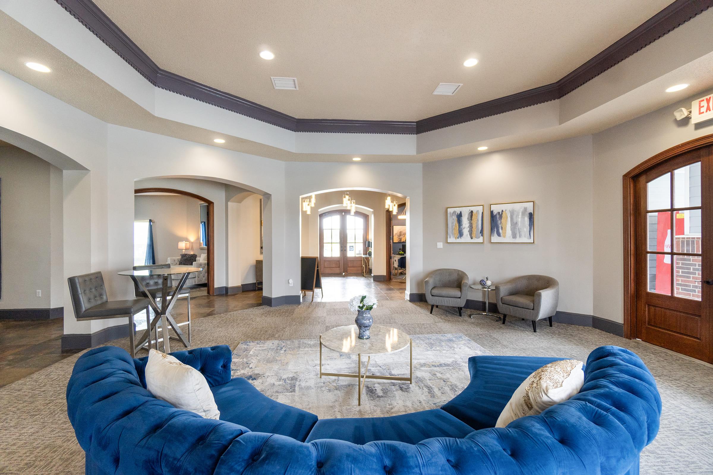 A spacious, modern lobby featuring a blue velvet sofa, two gray chairs, a coffee table with a flower centerpiece, and abstract artwork on the walls. The ceiling has a subtle architectural detail, and there are warm wood doors in the background leading to another area. Natural light fills the space through large windows.