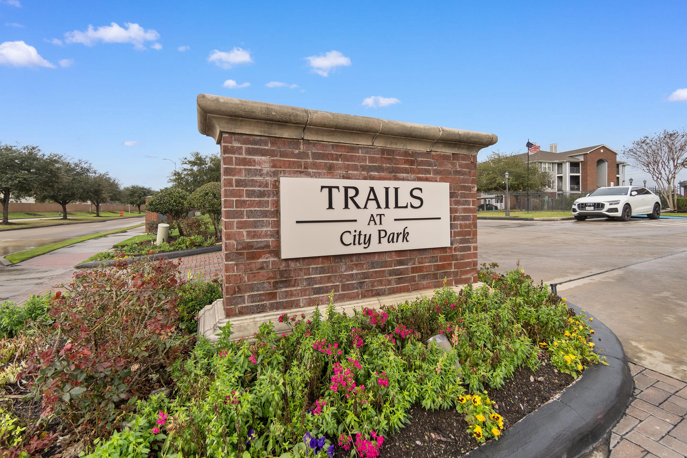 Sign reading "Trails at City Park" surrounded by vibrant flower beds. In the background, a residential building and a car parked nearby. Clear blue sky adds to the welcoming atmosphere of the property entrance.