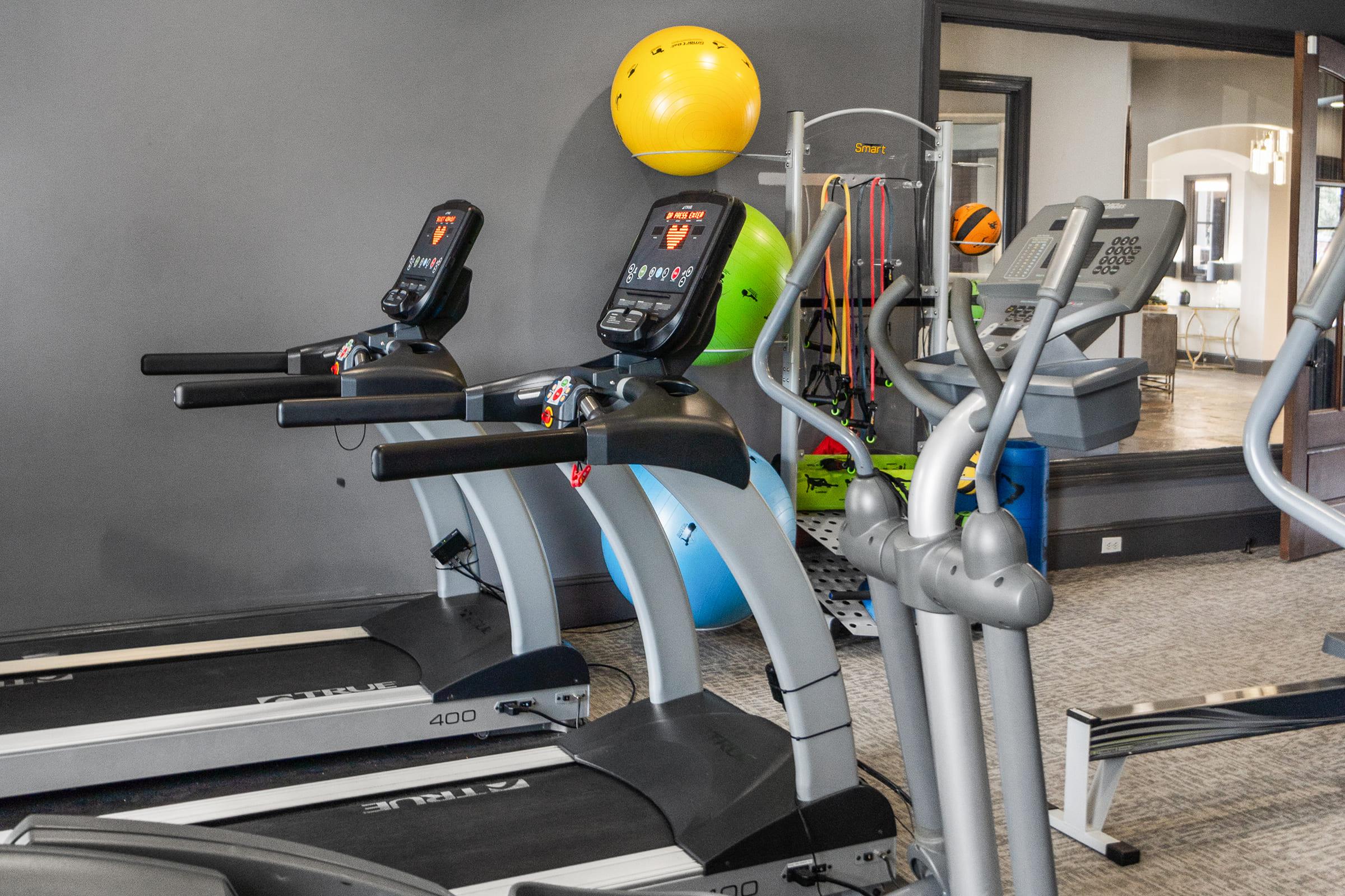 A fitness center featuring several exercise machines, including treadmills with digital displays and an elliptical. In the background, there are stability balls and resistance bands on a rack, along with other gym equipment. The walls are painted gray, creating a modern workout environment.