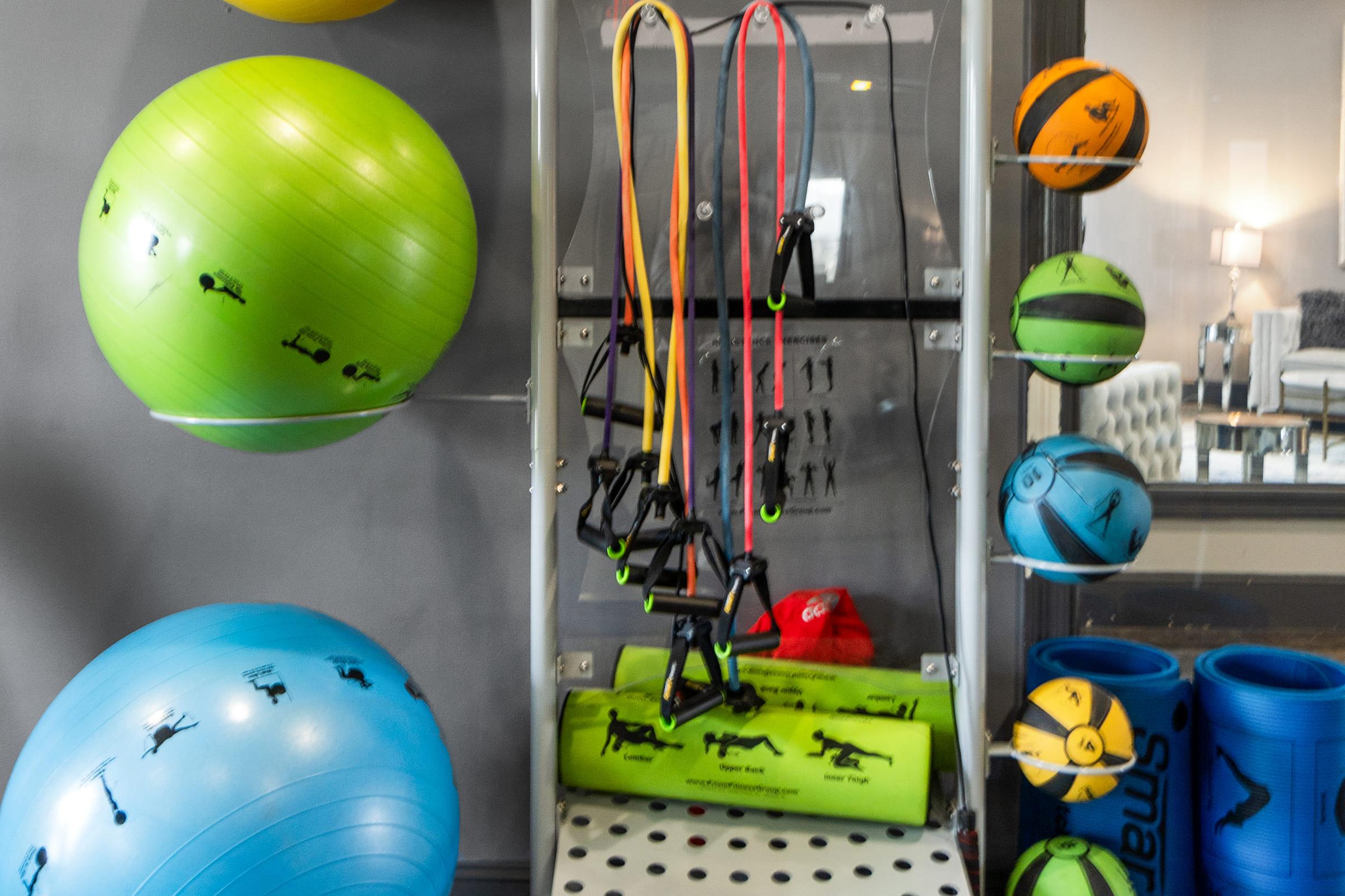 A variety of fitness equipment, including green and blue exercise balls, resistance bands in yellow, red, and orange, a green foam roller, and various colorful Pilates rings displayed on a shelf. The background features blurred furniture and a light fixture.