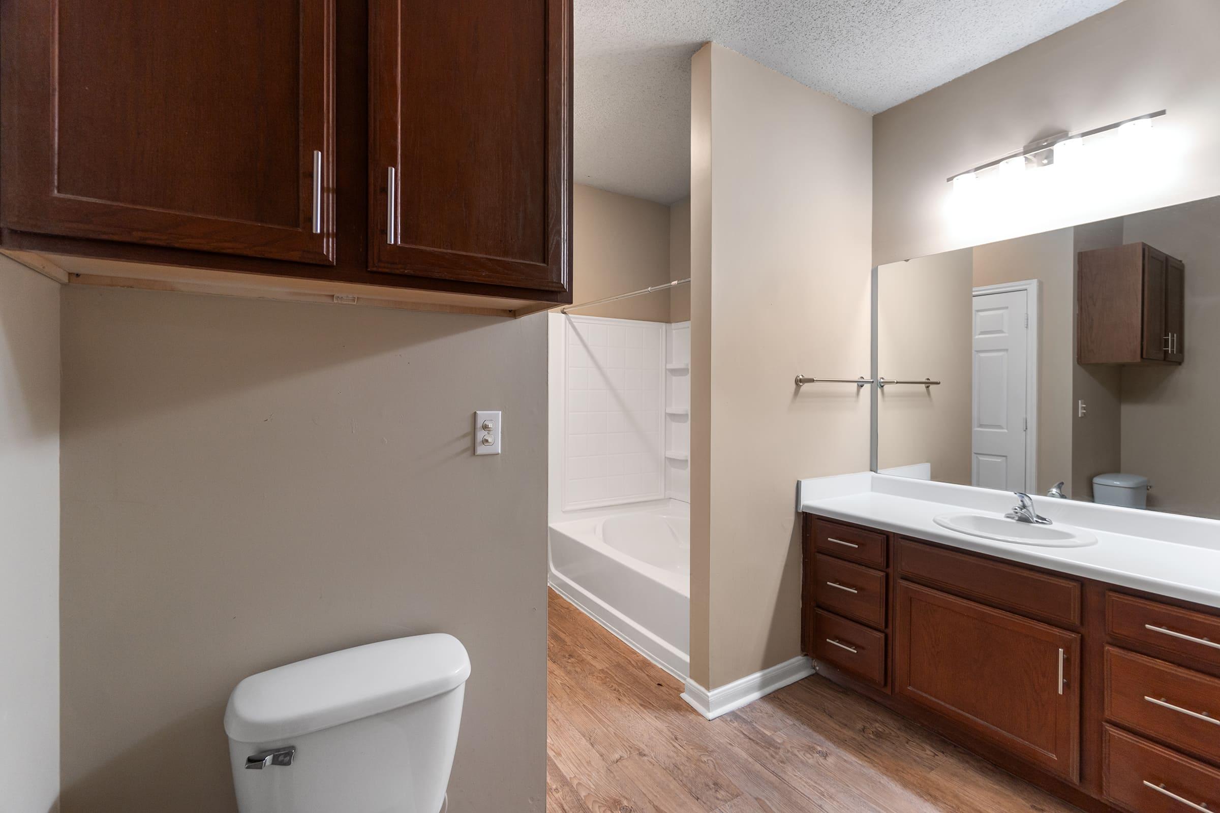 A modern bathroom featuring a white bathtub, a large mirror above a double sink vanity with dark wood cabinets, and a light-colored wall. There’s a toilet visible, along with a shower area. The flooring appears to be wood-like laminate, giving the space a contemporary look.