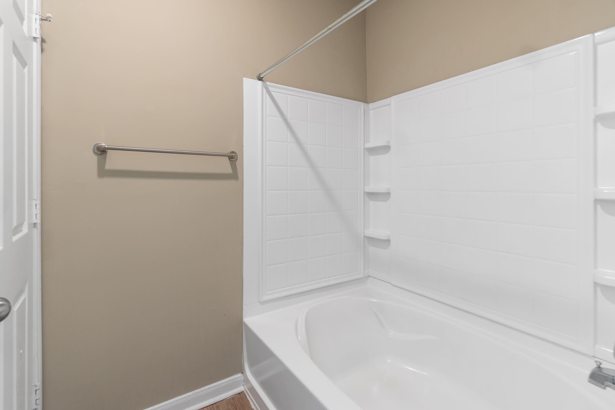 A clean bathroom featuring a white bathtub with a shower rod and a tiled wall. The walls are painted in a neutral beige color. A towel rack is mounted on the wall, and the flooring appears to be light wood. The overall ambiance is simple and functional.