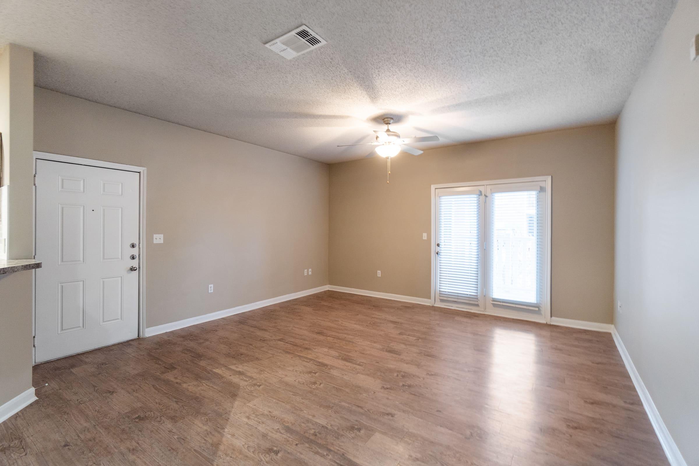 A spacious, empty living room featuring light brown walls and laminate flooring. There's a ceiling fan and a pair of glass sliding doors leading to an outdoor area. A front door is visible on the left, and the overall ambiance is bright and open.
