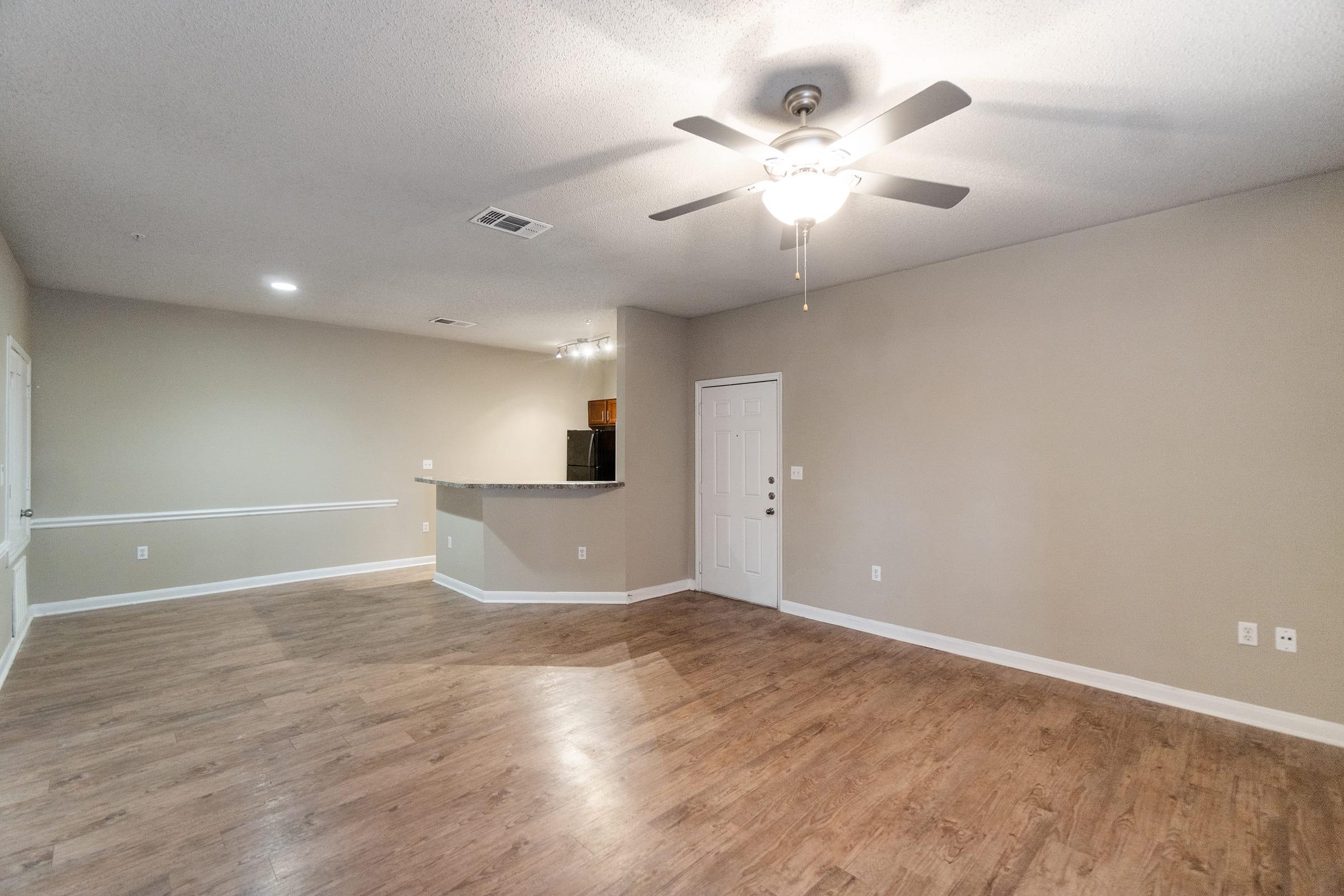 A spacious, unfurnished living room with light-colored walls and hardwood flooring. It features a ceiling fan, a doorway leading to another room, and an open area that connects to a kitchen with a bar counter in the background. Natural light is coming from a nearby window.