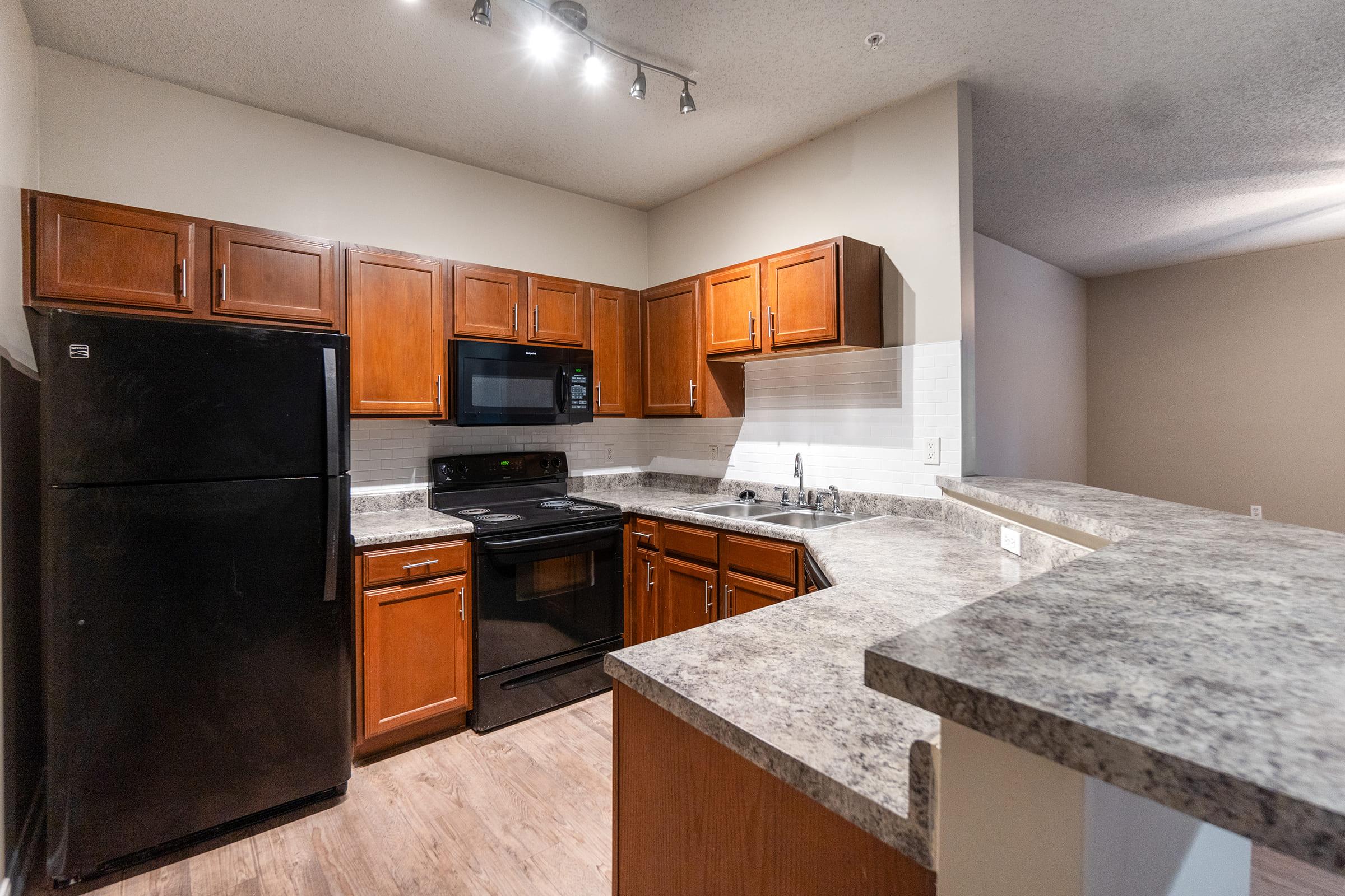 Modern kitchen featuring wooden cabinets, stainless steel appliances including a refrigerator and microwave, and a granite countertop. The space is well-lit with overhead lighting, showcasing a clean and organized layout. The floor is covered in light-colored wood laminate.