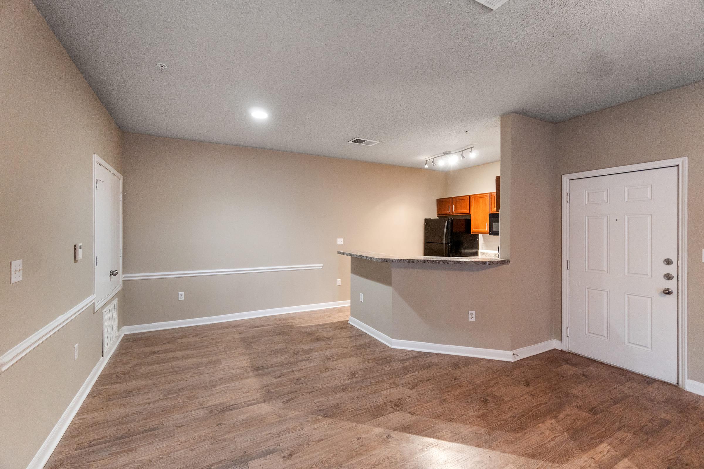 A spacious, unfurnished apartment interior featuring a neutral-colored wall and hardwood flooring. The room includes a partially open kitchen with dark cabinets and appliances visible in the background. There is a front door on the right, and light fixtures on the ceiling provide illumination.