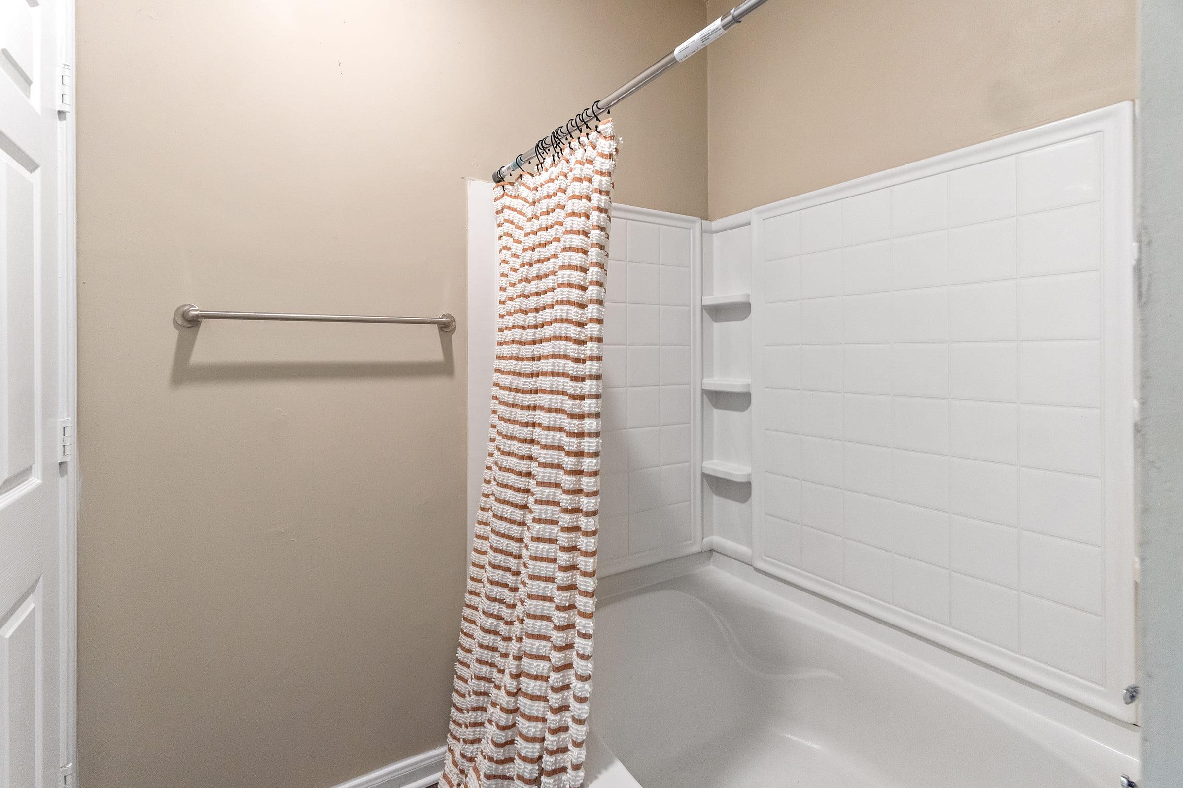A bathroom featuring a bathtub with a shower curtain, a towel rack on the wall, and white tiled walls. The color scheme is neutral, with light brown walls and a clean, modern design. The shower curtain has an orange and white pattern, adding a pop of color to the space.