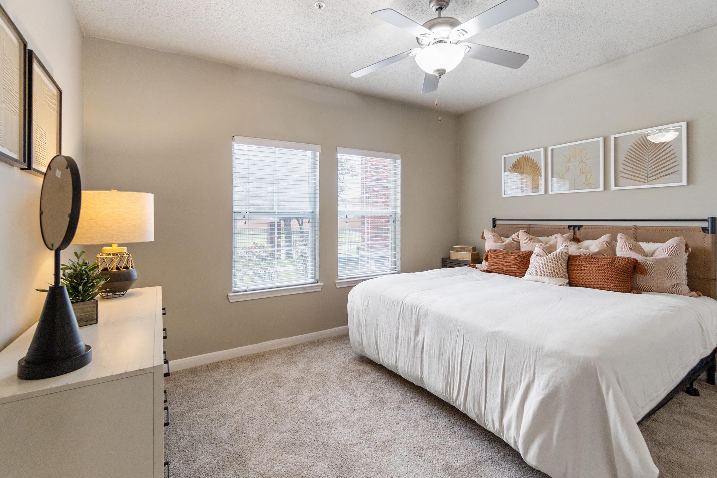 Cozy bedroom featuring a large bed with decorative pillows, light-colored bedding, and a stylish headboard. Natural light streams through two windows, illuminating the space. A bedside lamp and a small plant add charm, while framed artwork adorns the walls. The room has a soft, neutral color palette.