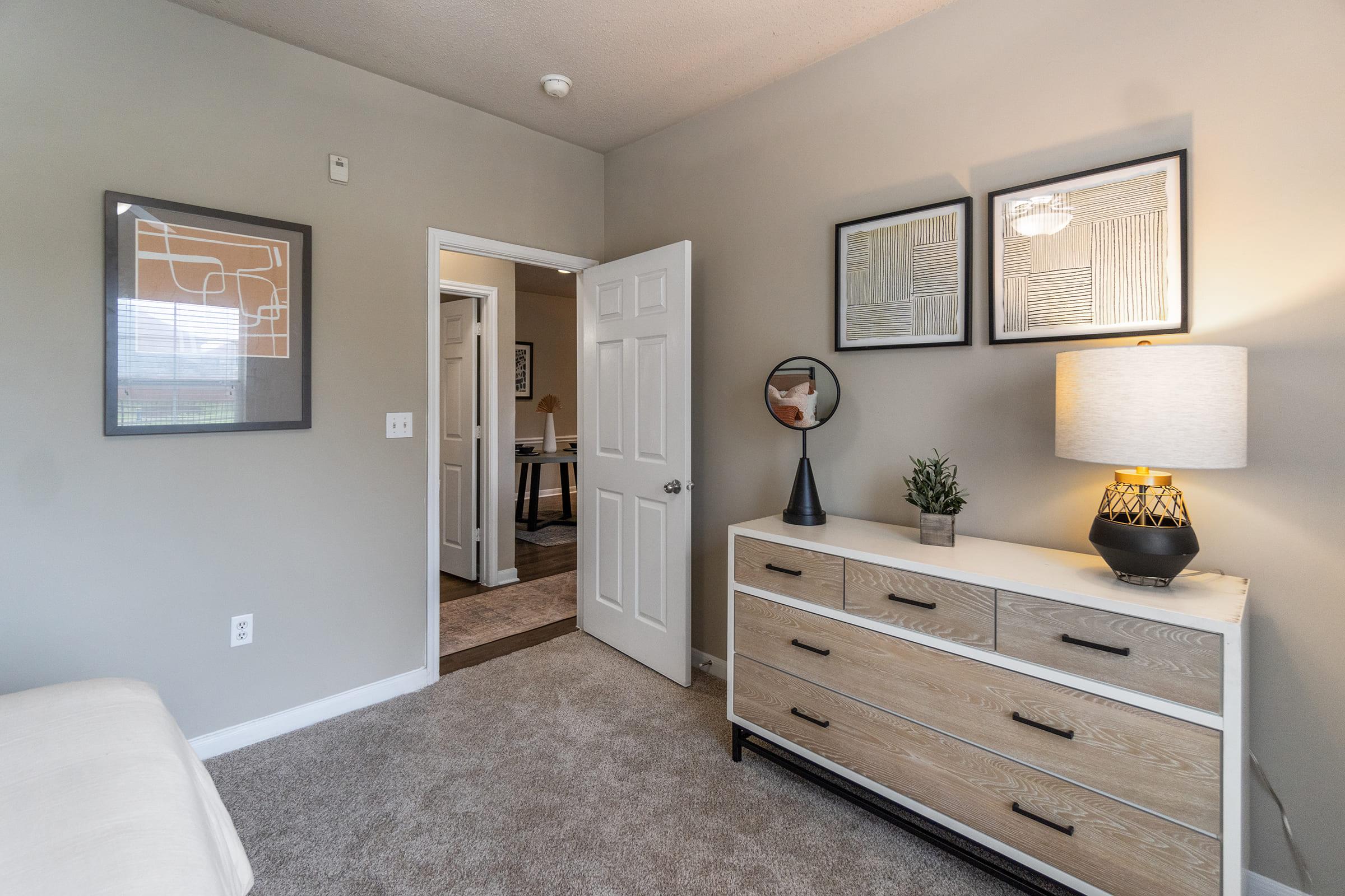 A cozy room featuring a light-colored dresser with drawers, a decorative lamp, and framed wall art. The walls are painted a soft gray, and there is a visible door leading to another room in the background. Neutral carpeting adds warmth to the space.