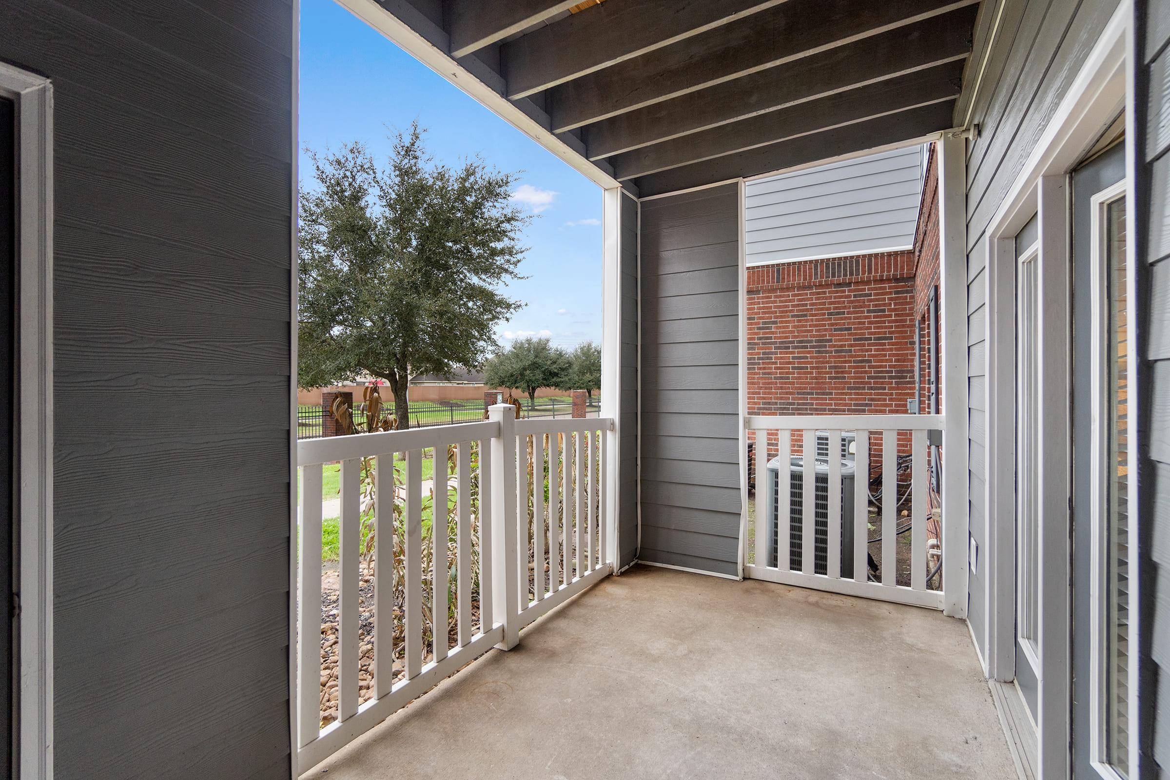View of a small balcony or porch with a concrete floor and white railings. The surrounding area features trees and grassy space, with a clear blue sky above. The adjacent buildings have a modern design, showcasing a blend of wood and brick exteriors.